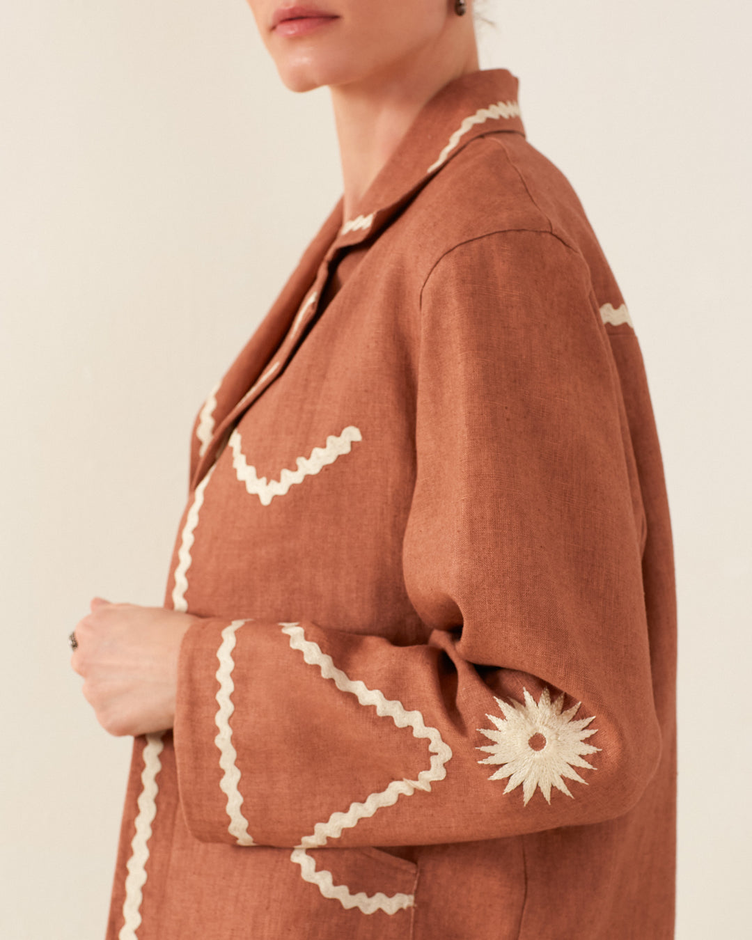 A person is dressed in the Western Clove Ric Rac Jacket, which showcases white embroidered patterns on its sleeves and collar. With a sunburst design on the elbow and zigzag accents reminiscent of a retro floral cotton waistcoat, only their lower face and torso are visible against a plain background.