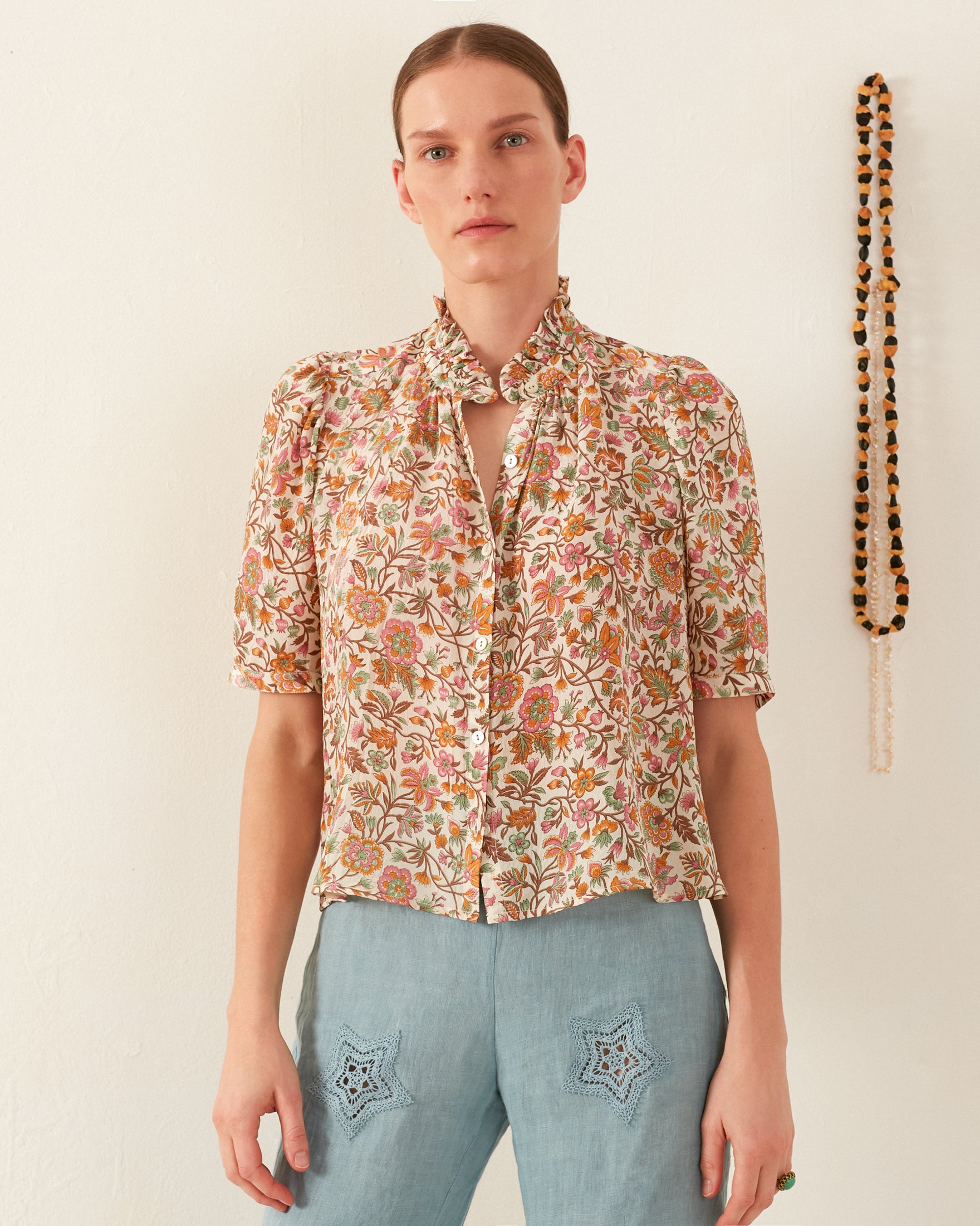 A person in the Winn Garden Party Silk Shirt and light blue pants stands against a plain wall. They have a neutral expression, and a heavyweight beaded necklace hangs on the wall to their right.