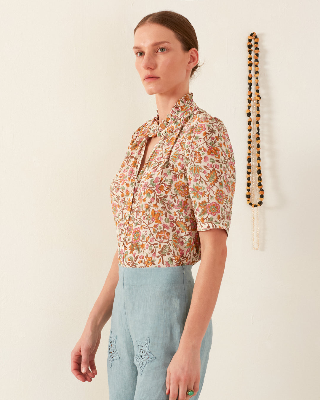 A person with hair tied back is wearing a Winn Garden Party Silk Shirt and light blue pants. They stand against a plain wall with a beaded necklace hanging in the background.