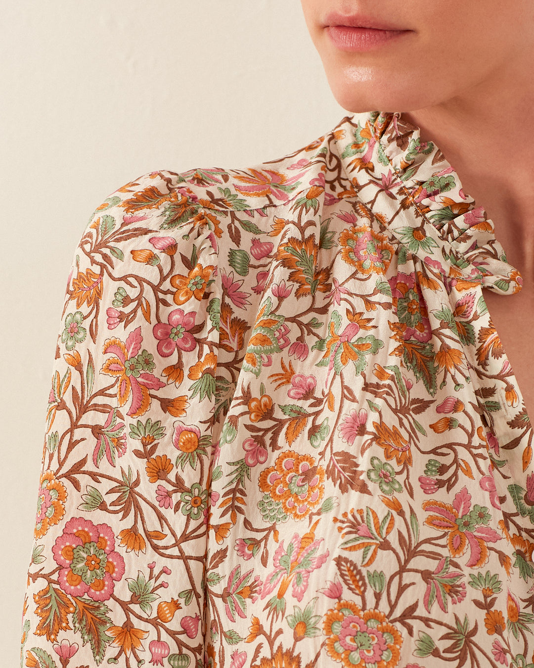 A person wearing the Winn Garden Party Silk Shirt, which boasts a floral pattern in pink, orange, and green on a light background. The shirt features a ruffled collar, and the individual's lips are visible against the plain, neutral background that enhances the delicate fabric.