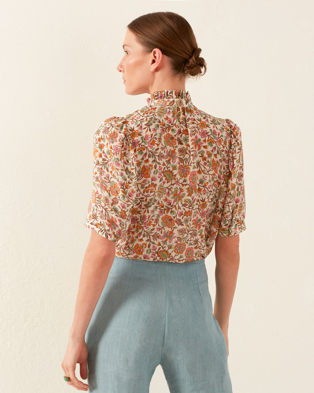 A person with short hair is wearing the Winn Garden Party Silk Shirt, featuring a high collar and puff sleeves, crafted from silk cotton voile with a floral design. They have paired it with light blue pants and are standing against a plain light-colored background, turned away while looking off to the side.