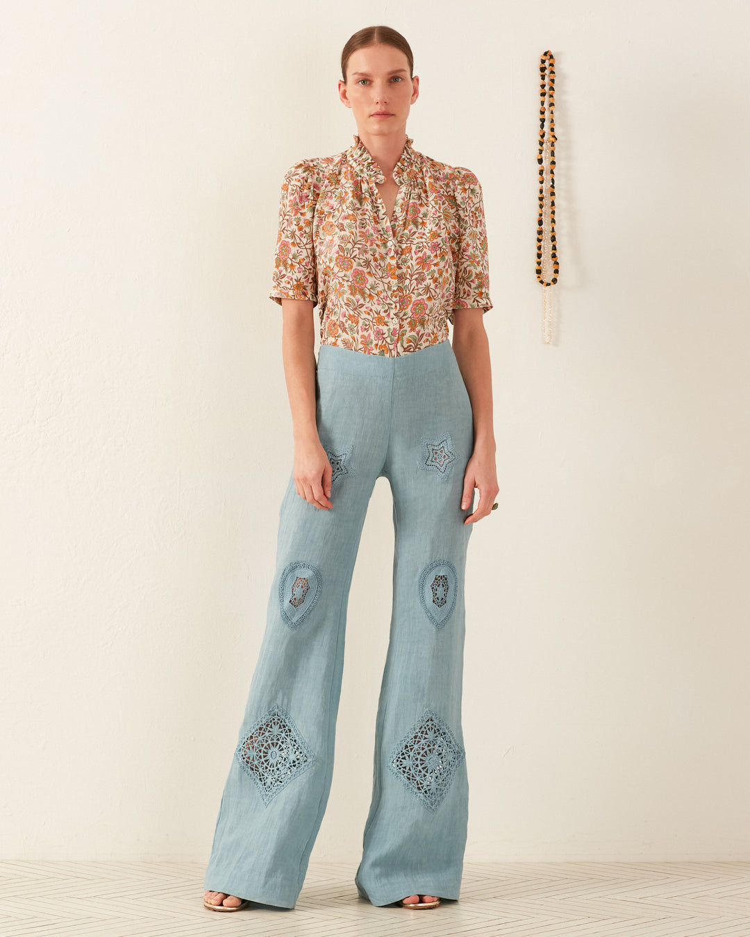 A person stands against a plain wall, wearing the Winn Garden Party Silk Shirt adorned with a floral design and high-waisted, light blue pants featuring diamond-shaped embroidery. A beaded necklace hangs on the wall beside them.