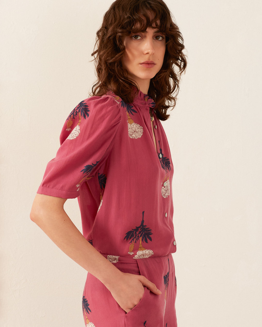 A person with wavy brown hair is wearing the vintage-inspired Winn Hibiscus Rose Shirt paired with pink pants. They are standing against a plain light-colored background, looking towards the camera with a neutral expression, and have one hand in their pocket.