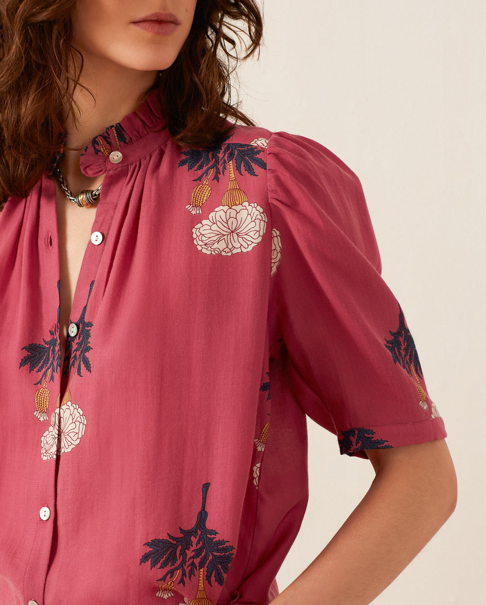 A woman wearing the Winn Hibiscus Rose Shirt, a vintage-inspired, mauve blouse adorned with a ruffled collar and floral print. This silk chiffon shirt is designed with buttons down the front and short sleeves. Her hair is loose and wavy.