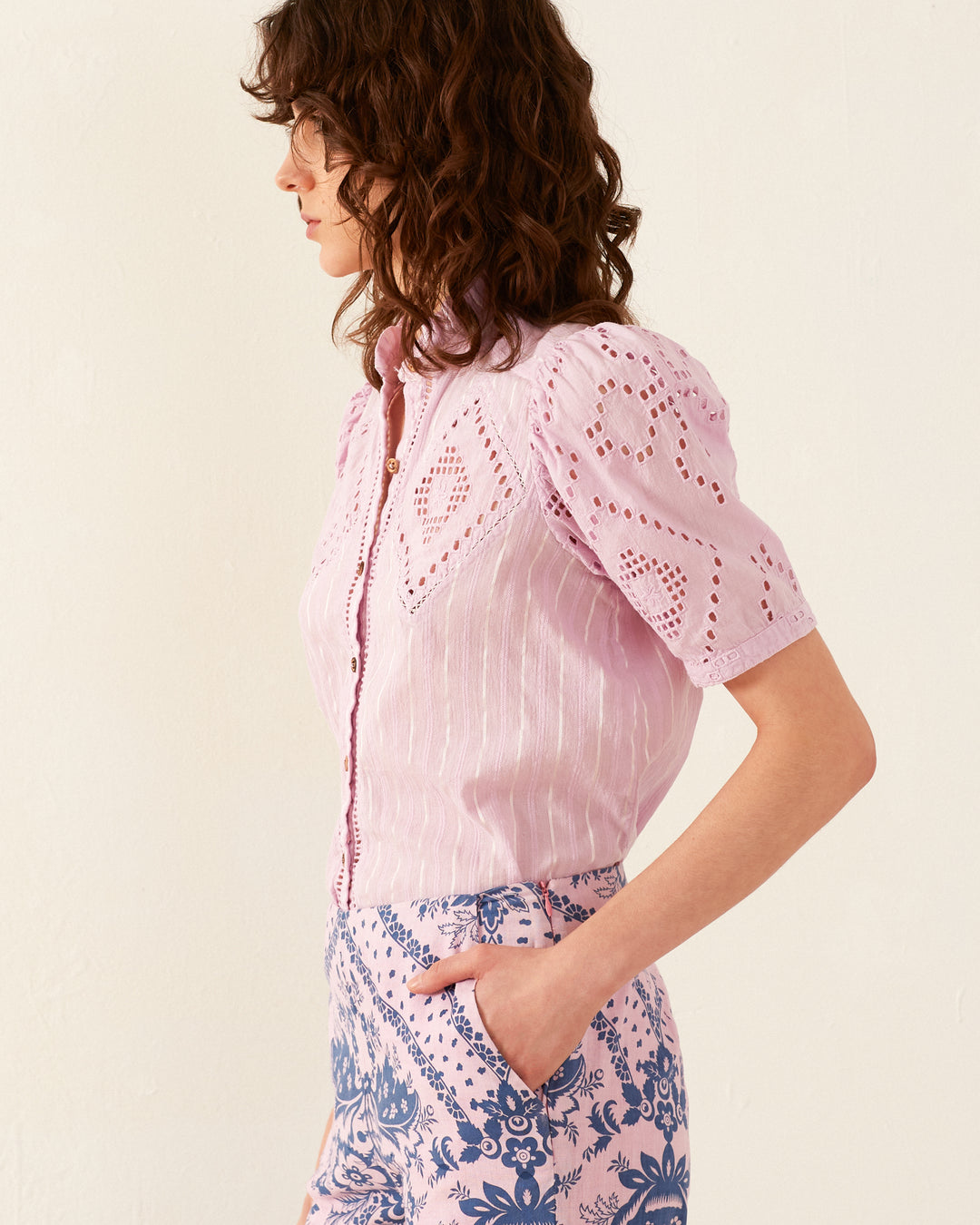 A person with wavy brown hair stands in profile against a plain wall, dressed in the Winn Lilac Eyelet Shirt reminiscent of '70s vintage-inspired style and high-waisted shorts adorned with a blue floral design. Their hand rests casually in their pocket.