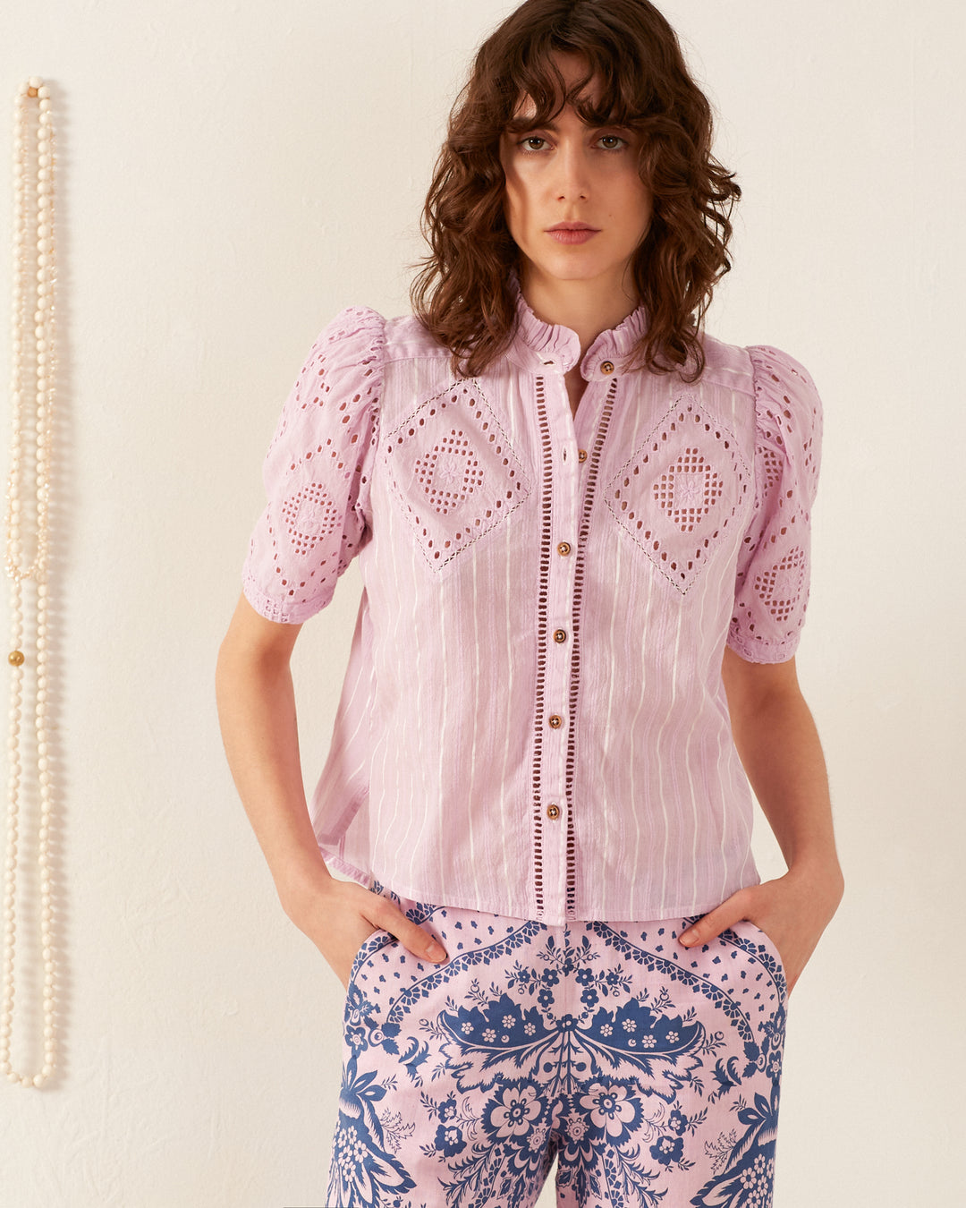 A person with shoulder-length curly hair wears the Winn Lilac Eyelet Shirt, featuring intricate cut-out designs and short sleeves, paired with '70s vintage-inspired patterned blue and white pants. They stand against a pale background, adorned by a strand of pearls hanging on the side.