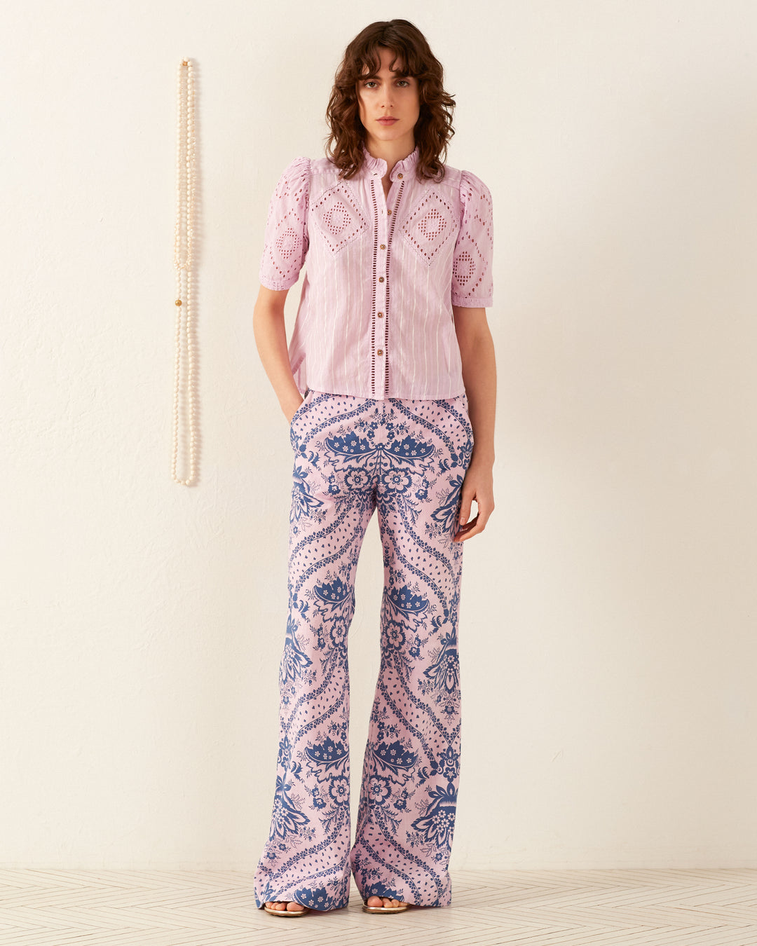 A person stands against an off-white wall adorned with hanging beads, wearing a short-sleeved '70s vintage-inspired Winn Lilac Eyelet Shirt and high-waisted, wide-leg pants featuring a hand-block printed pink and blue floral pattern. They have shoulder-length wavy hair.