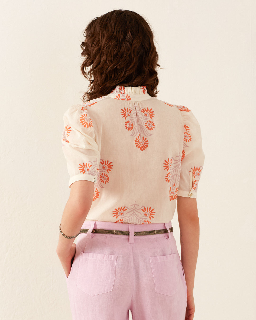 Facing away, a person with curly brown hair is wearing the Winn Papaya Aster Shirt, which features vintage-inspired orange floral patterns. They pair it with light pink pants and have their hands in their pockets, standing against a plain, off-white background.