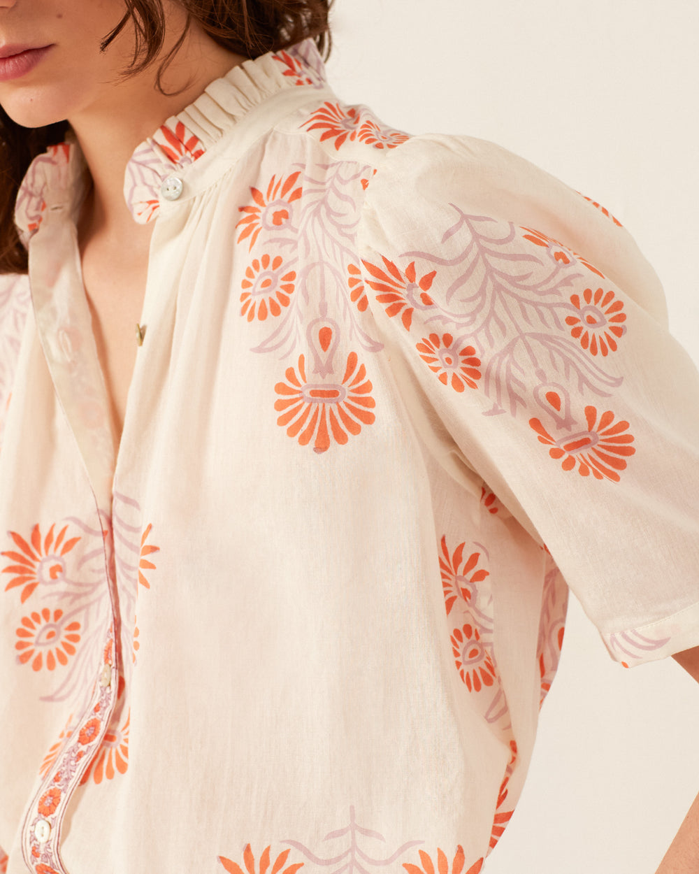 Close-up of a woman wearing the Winn Papaya Aster Shirt, a vintage-inspired summer top adorned with a floral pattern in orange and pink hues. It features puffed sleeves and a ruffled collar against a soft cream background.