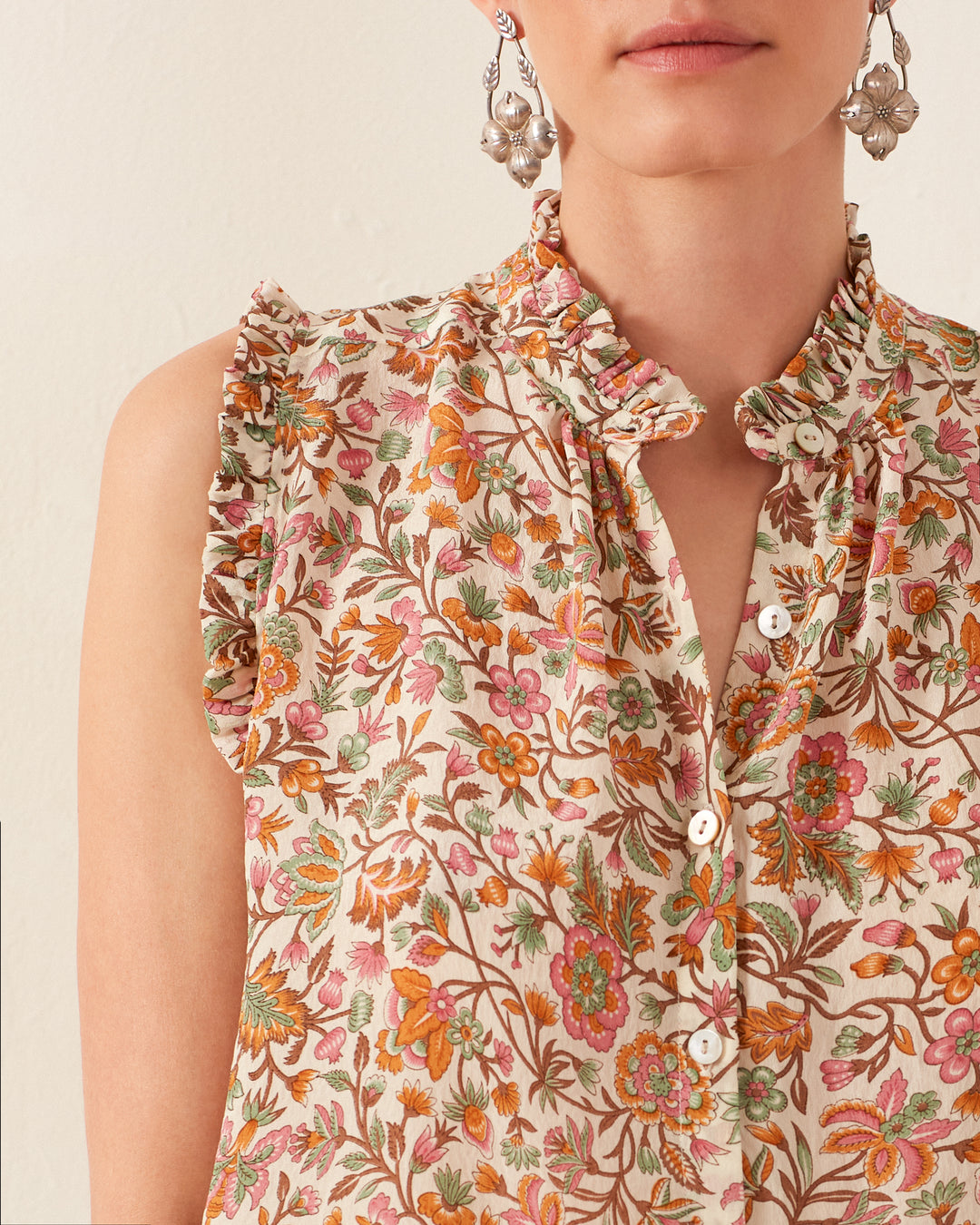 A person dons the Wylder Garden Party Silk Shirt, showcasing a colorful floral pattern in shades of orange, pink, and green. This sleeveless blouse features hand-loomed cotton fabric with ruffled edges at the sleeves and a buttoned neckline, perfectly paired with intricate silver floral earrings.