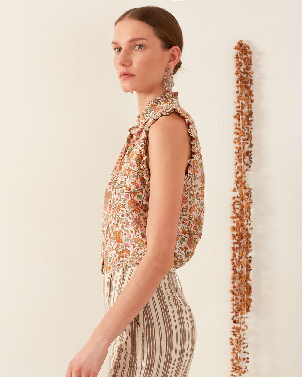 A person stands in profile against a neutral background, wearing the Wylder Garden Party Silk Shirt with its floral pattern and chic sleeveless design, paired with stylish striped pants. Their geometric-print shirt adds a modern twist. Dangling earrings complete the look, while a vertical row of dried flowers is visible behind them.