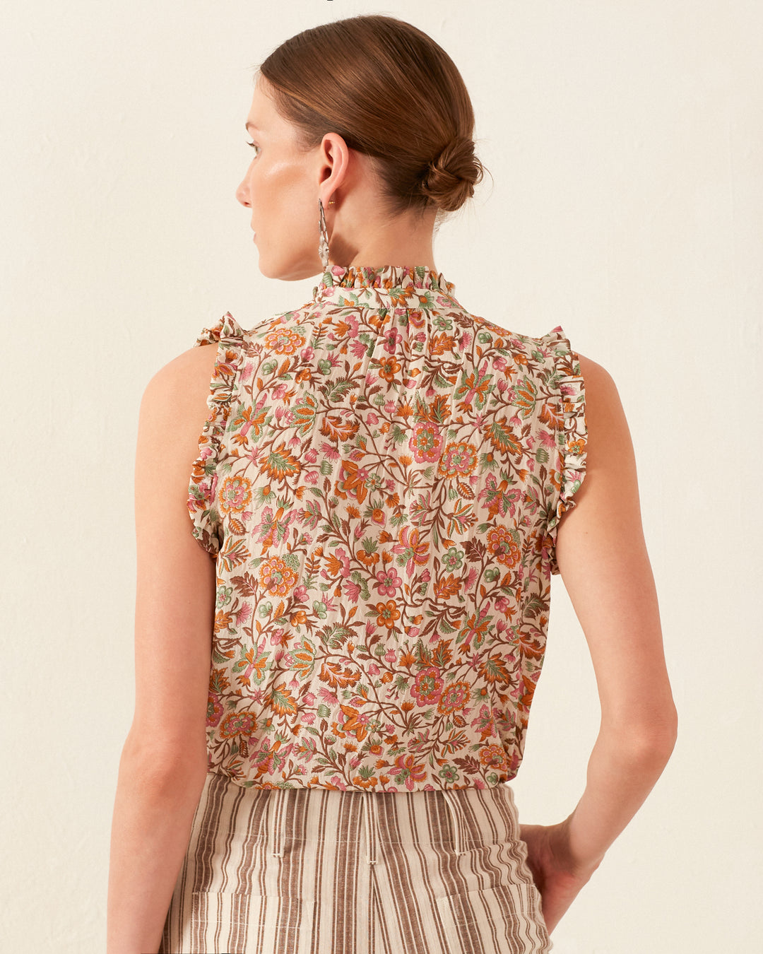 A woman with her hair tied back is facing away, wearing the Wylder Garden Party Silk Shirt, a sleeveless, floral top featuring ruffled shoulders. She pairs it with striped pants and large hoop earrings, all set against a plain, light-colored wall.