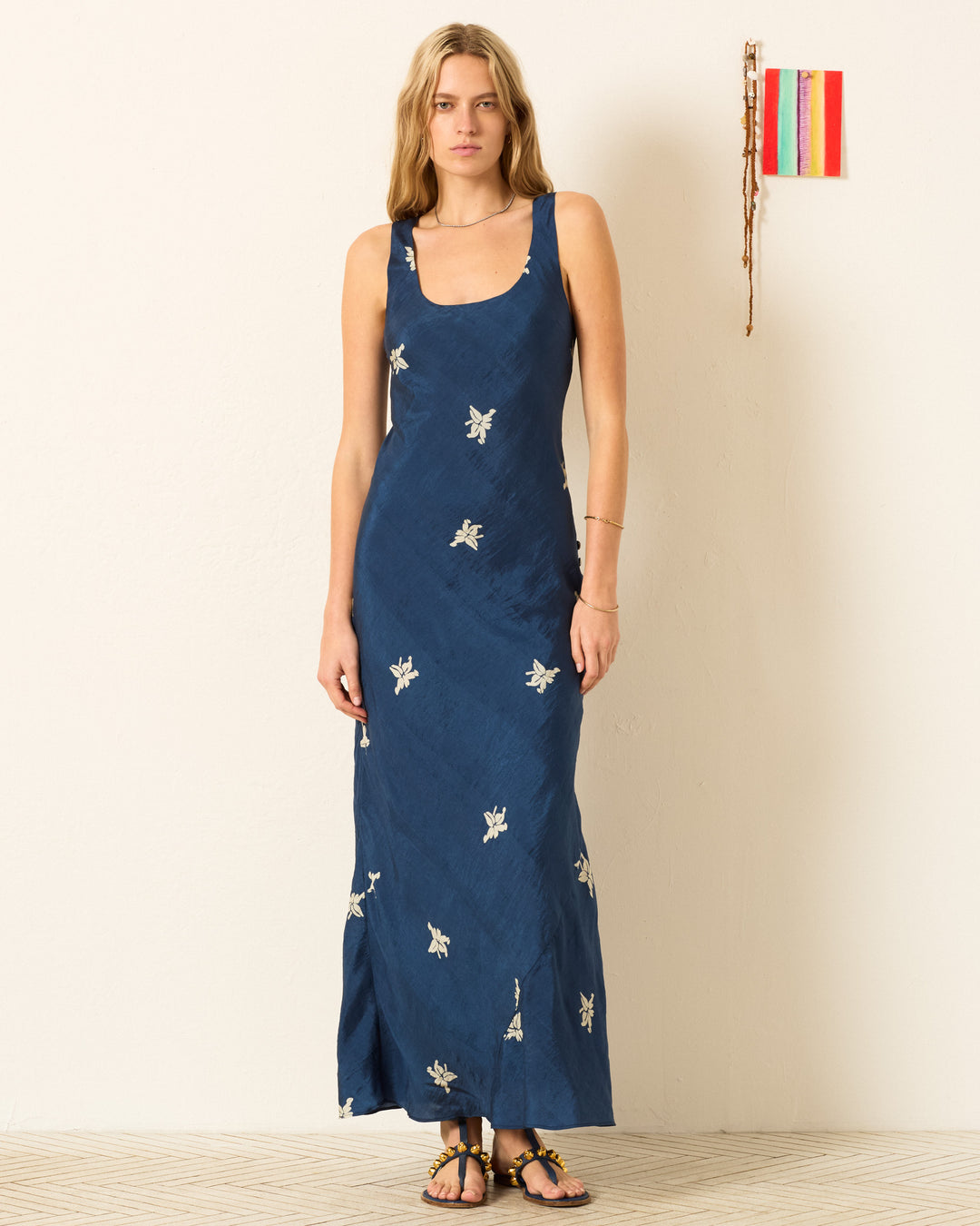 A woman stands against a plain wall wearing the Adele Pacific Jasmine Dress, a blue hand-block printed maxi with white floral patterns, paired with black sandals. A colorful striped decoration hangs on the wall to her right.