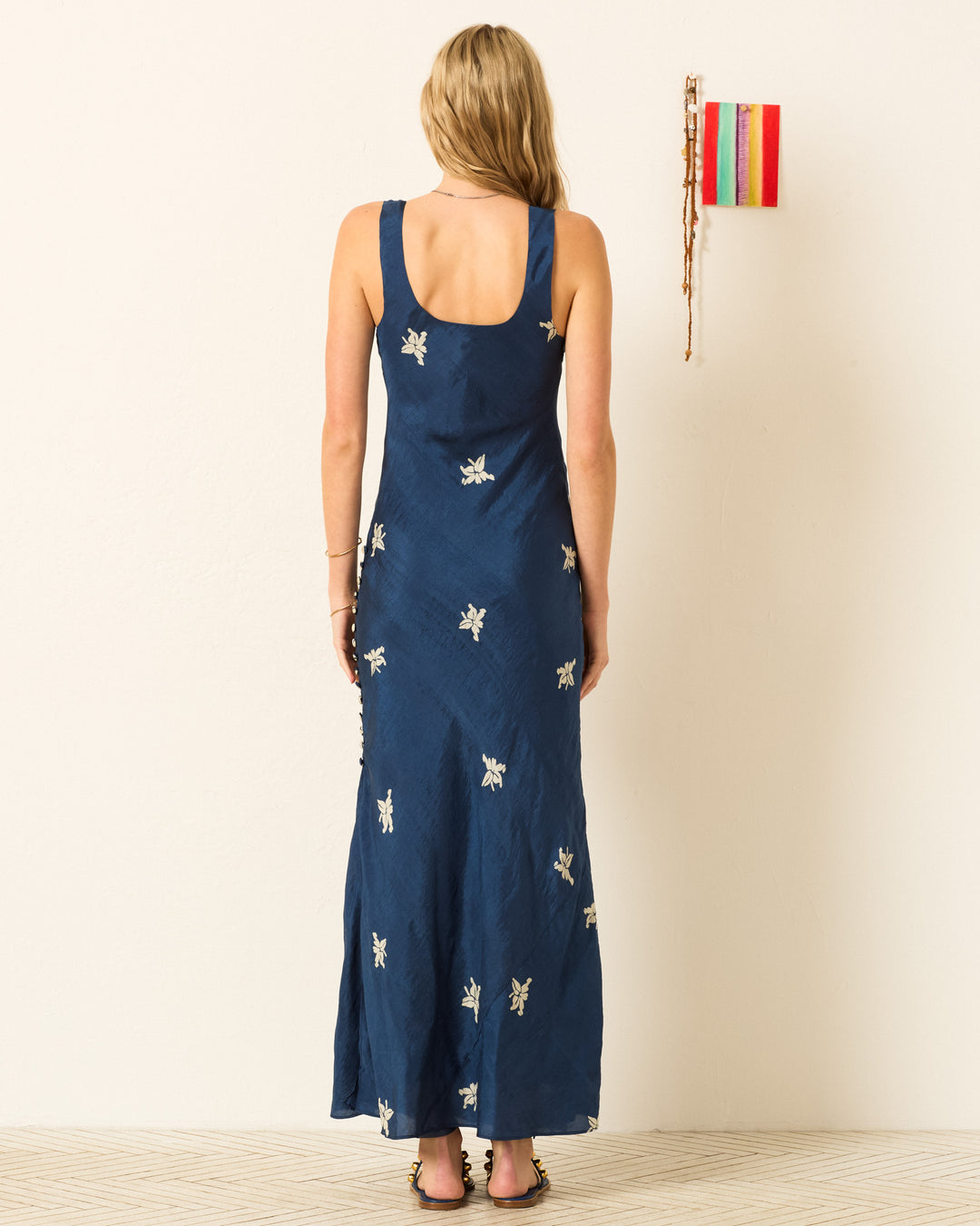 A woman with long blonde hair stands with her back to the camera against a plain wall, wearing the sleeveless navy Adele Pacific Jasmine Dress featuring white hand-block printed floral accents.