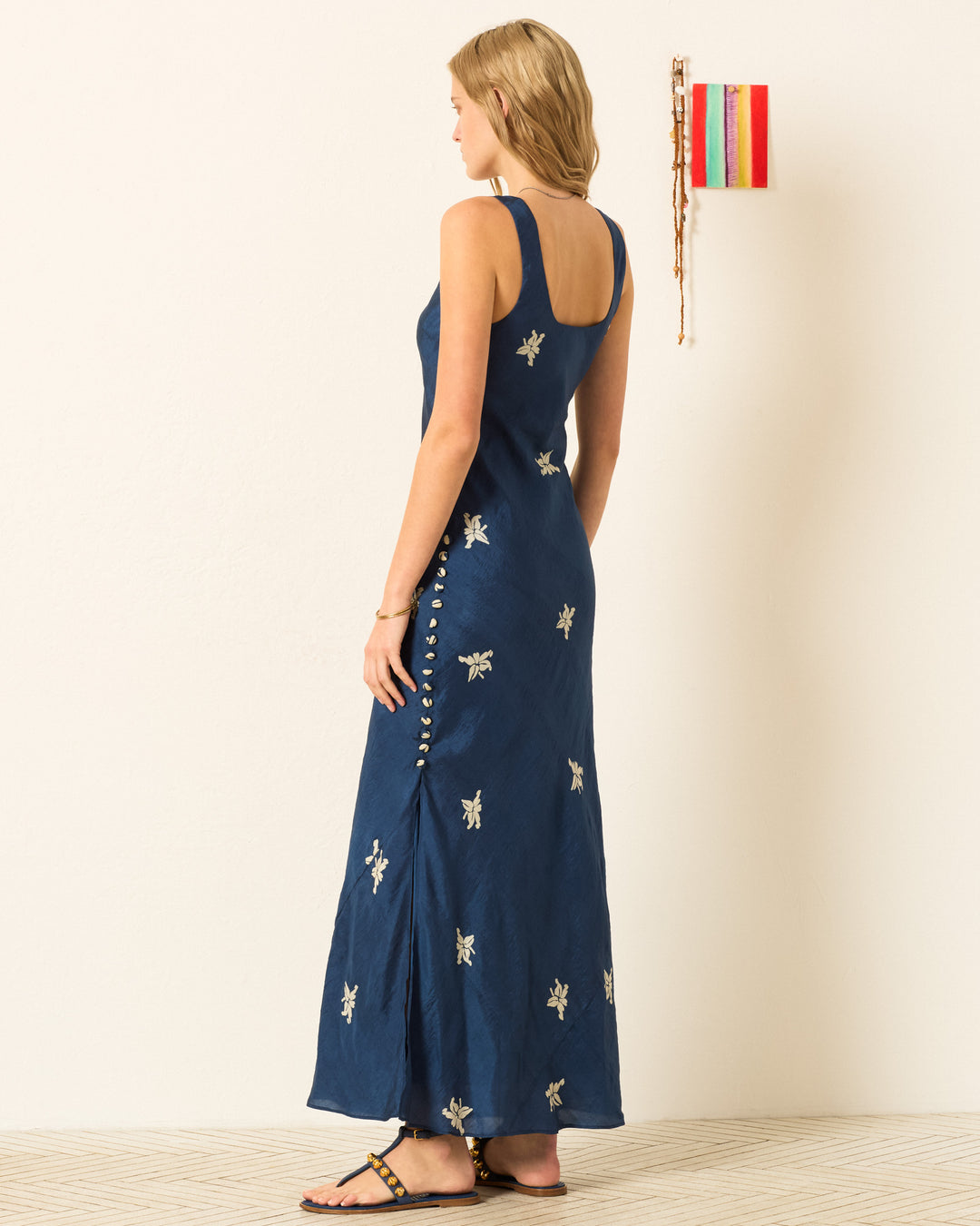 A woman stands sideways wearing the Adele Pacific Jasmine Dress, a sleeveless navy blue hand-block printed maxi with embroidered details and side button accents; simple wall decor appears in the background.