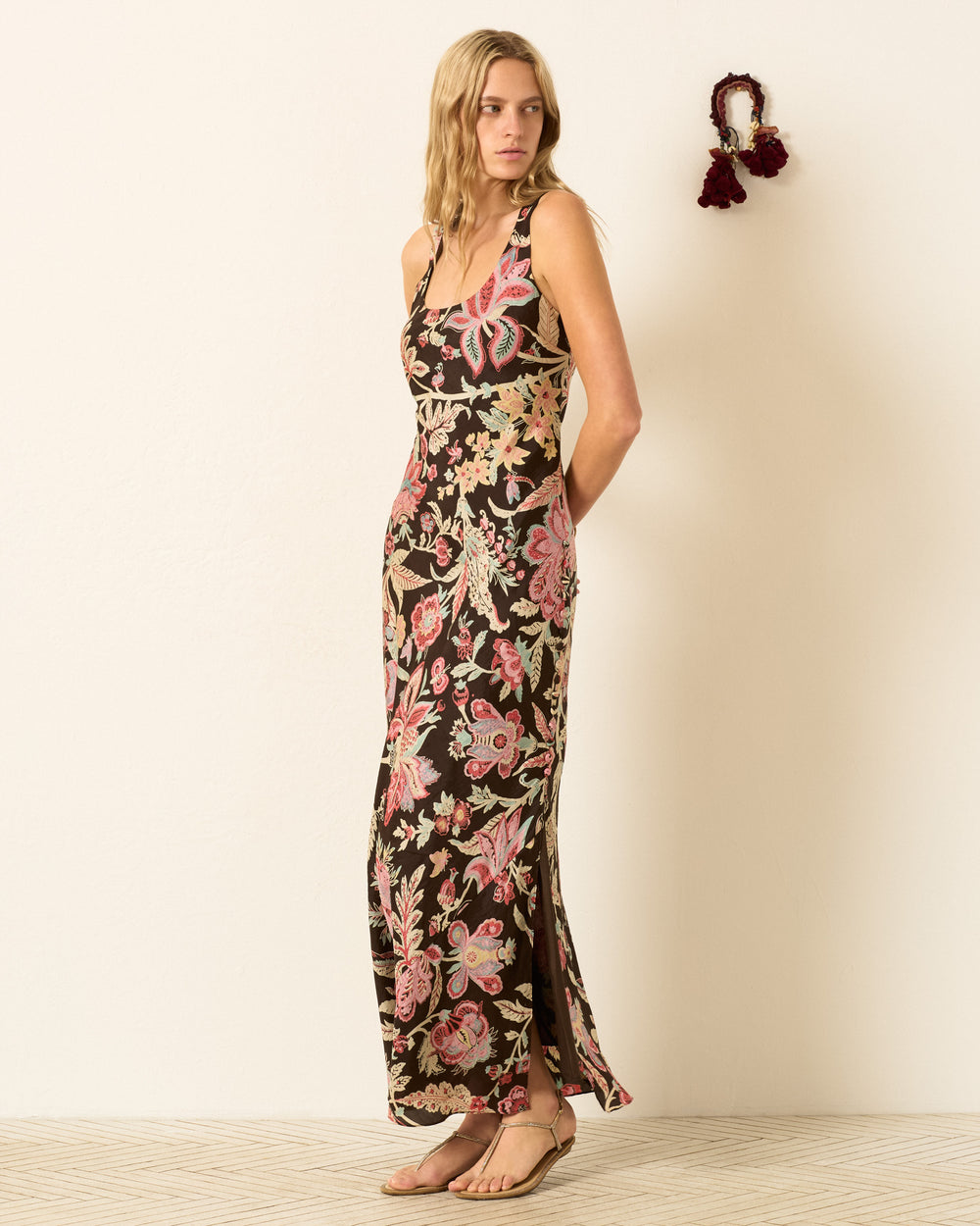 A woman stands against a light wall wearing the Adele Terra Floral Silk Dress, a sleeveless, ankle-length black maxi covered in pink and beige flowers, paired with flat sandals; a decorative tassel hangs nearby.