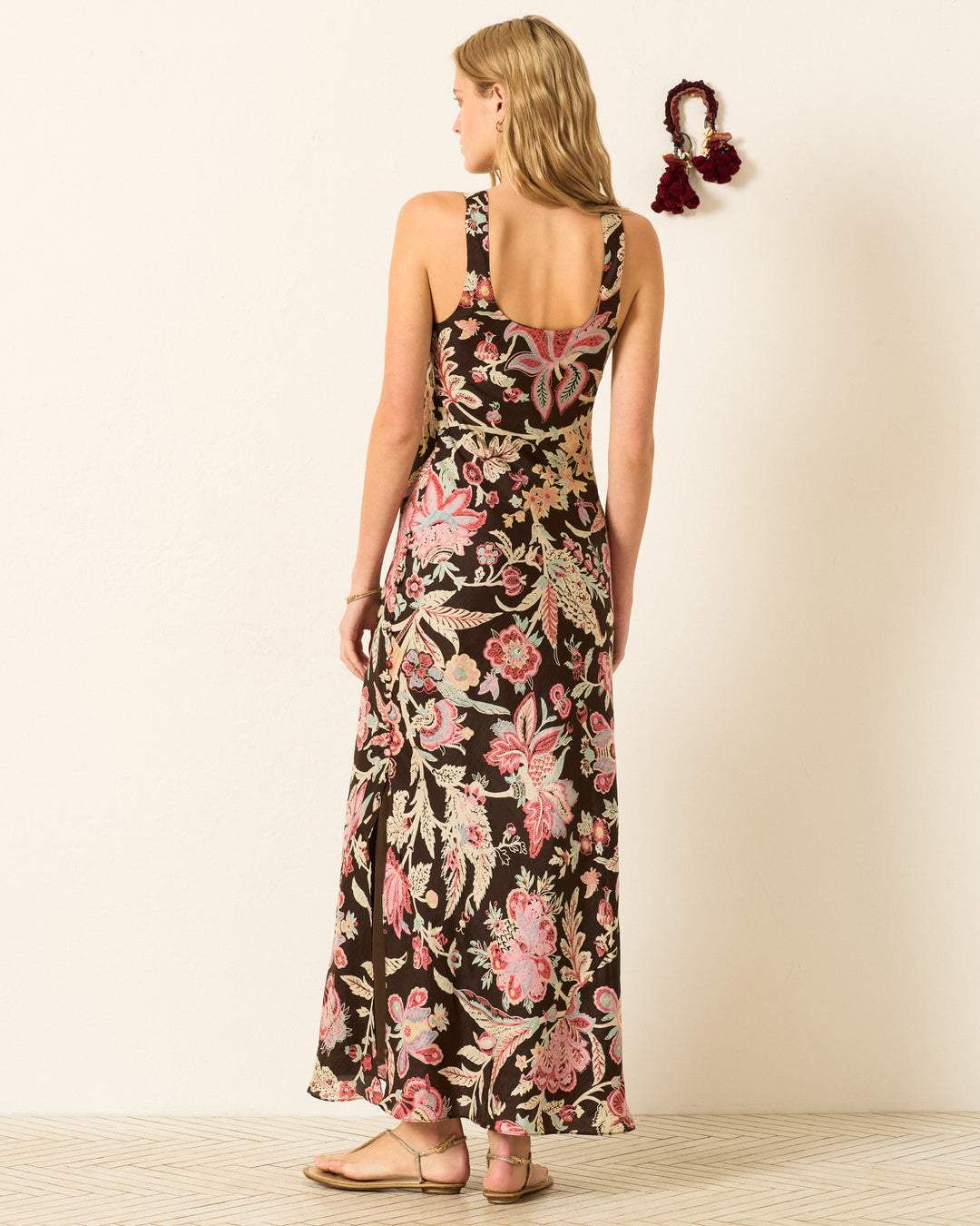 A woman stands facing away in the Adele Terra Floral Silk Dress with an open back and sandals, set against a plain wall featuring a decorative tassel.