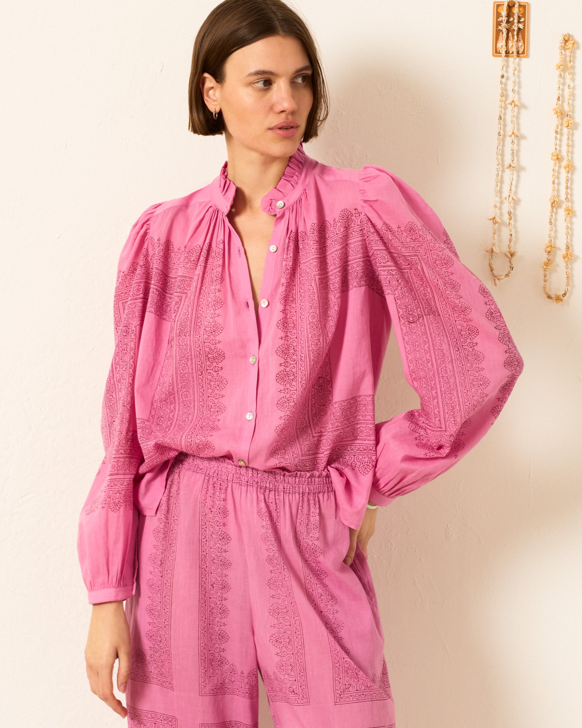 A woman wearing the Annabel Fuchsia Tile Shirt, a long-sleeved button-up blouse with puff shoulders and a high collar, paired with matching pink high-waisted pants, stands against a light wall decorated with hanging jewelry.