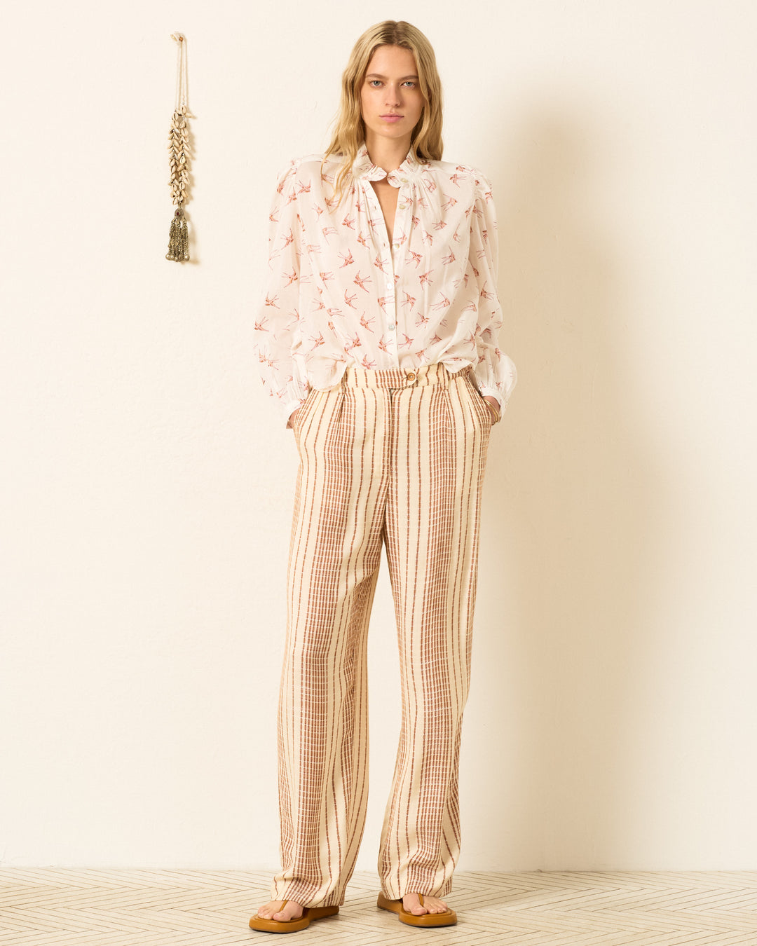 A woman stands indoors wearing the Annabel Cedar Swallow Shirt, a white hand-block-printed blouse with a bird motif, paired with beige striped pants. Her hands are in her pockets and a decorative tassel hangs on the wall behind her.
