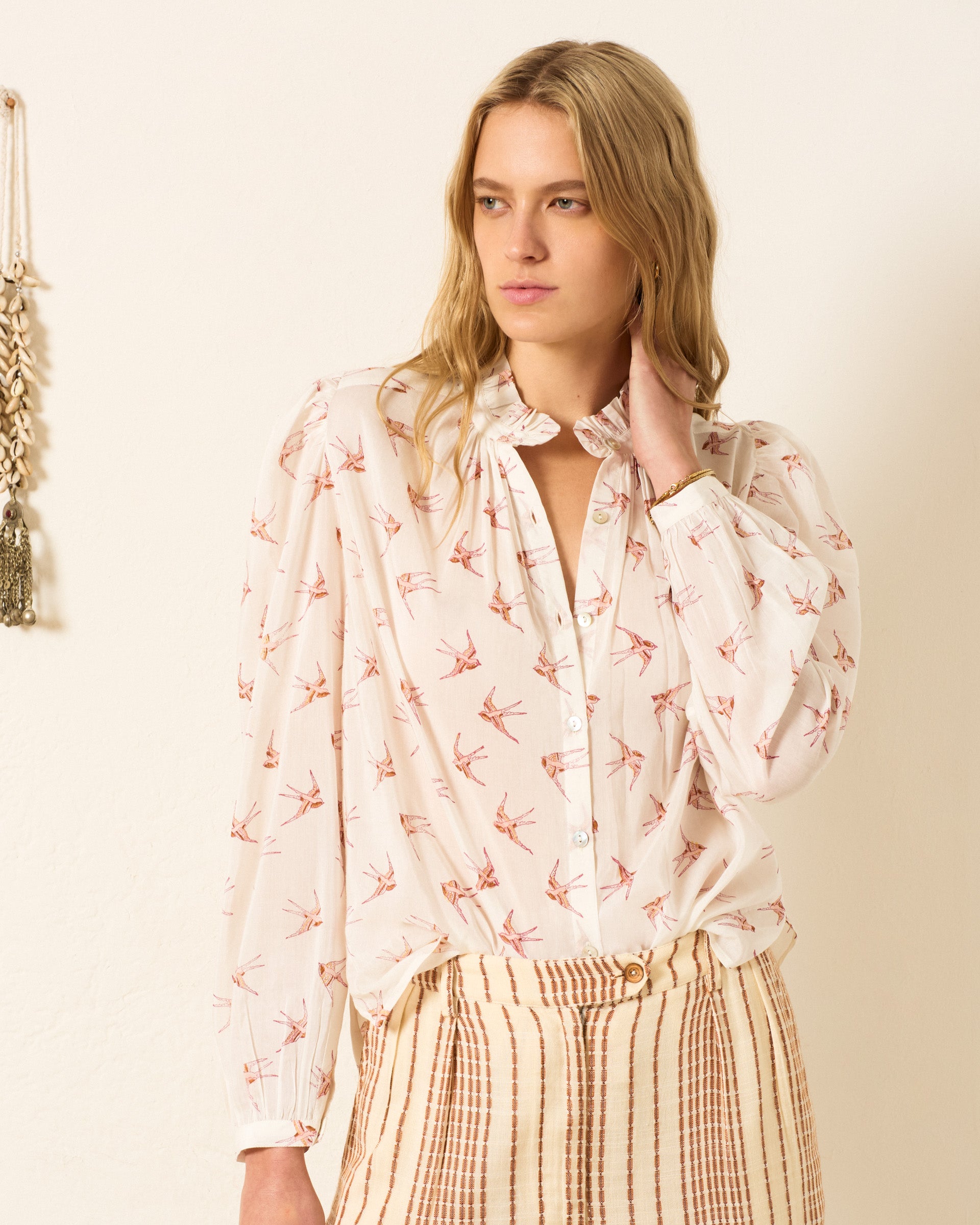 A woman stands indoors wearing the Annabel Cedar Swallow Shirt, a cotton-silk blend with a pink bird hand-block print, paired with beige striped pants and touching her neck.