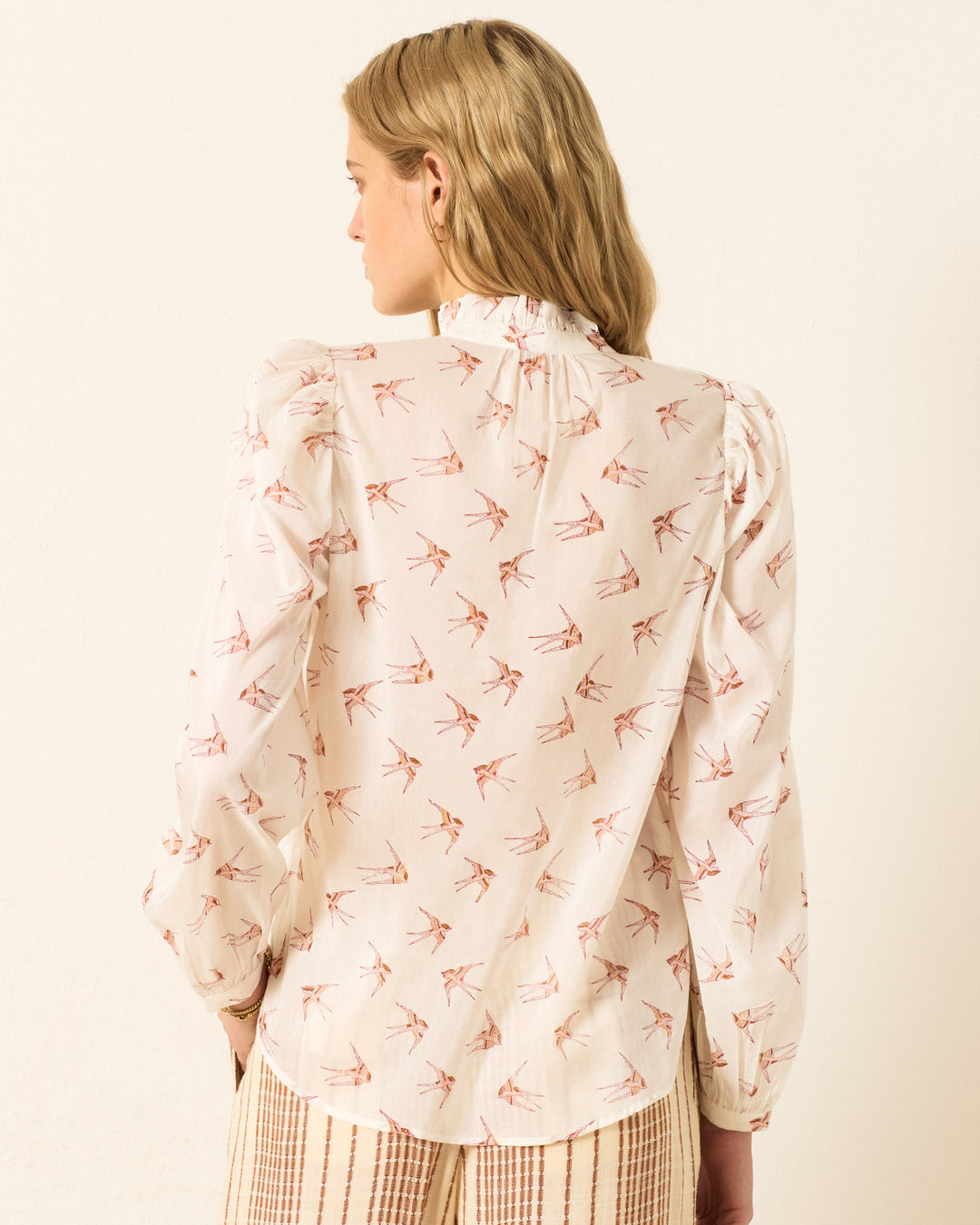 A woman with blonde hair is seen from behind, wearing the Annabel Cedar Swallow Shirt—a white, hand-block-printed, long-sleeve blouse with a bird motif—paired with light-colored striped pants.