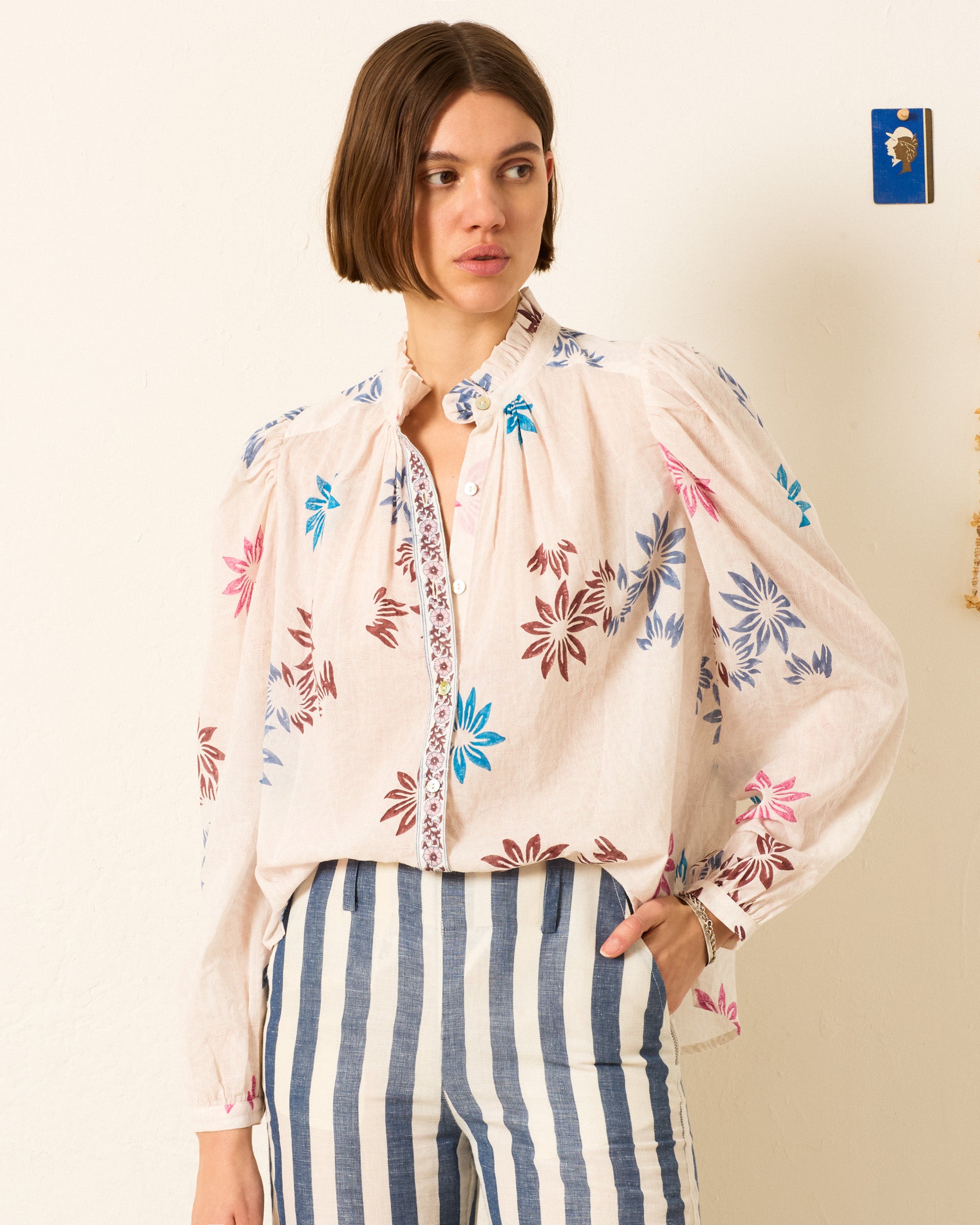 A woman wears the Annabel Berry Jackfruit Shirt, a light floral motif cotton voile top with puffed sleeves, paired with blue-and-white vertical striped pants, standing against a plain wall.