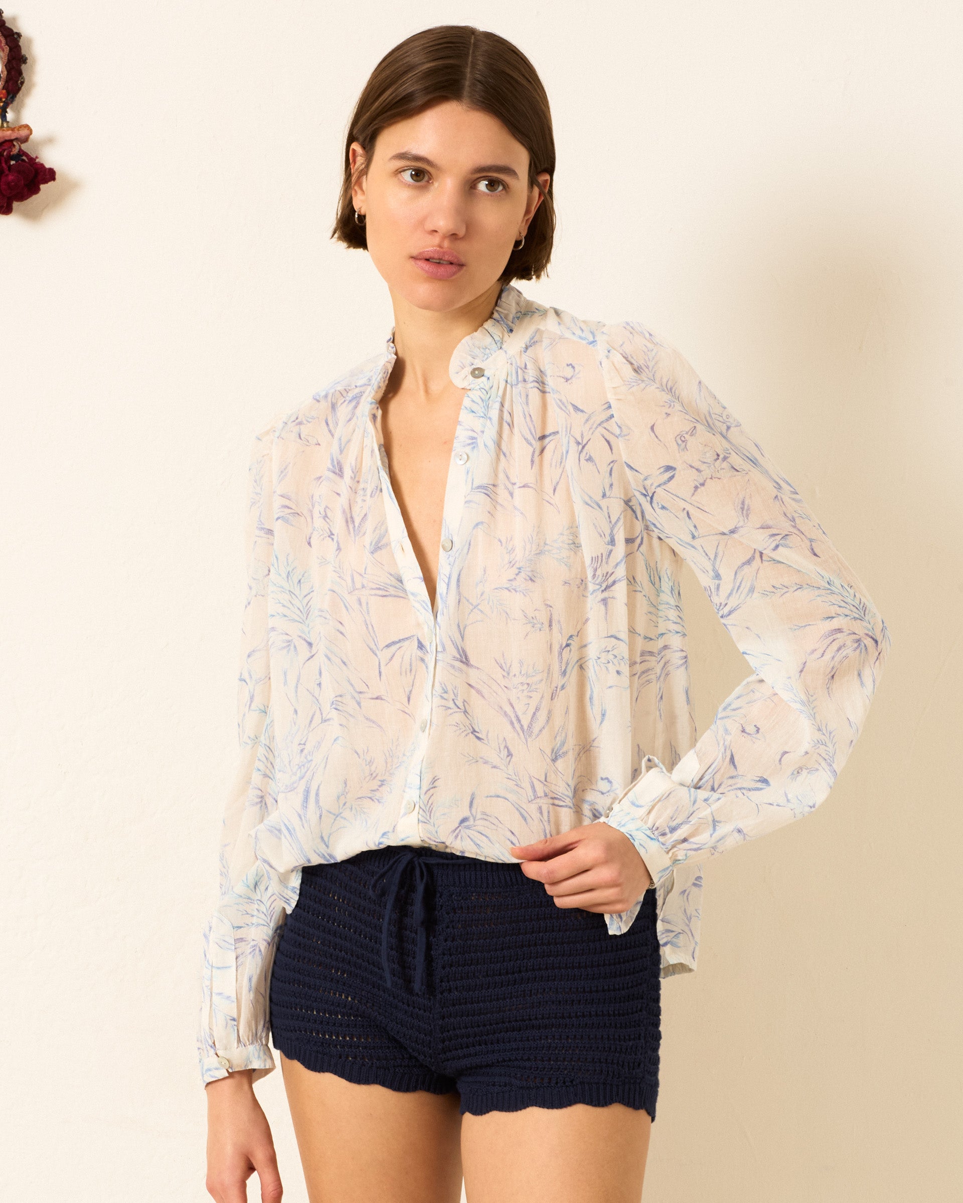 Woman wearing the Annabel Avian Toile Shirt—a sheer, long-sleeve floral cotton voile top with puff shoulders—paired with navy high-waisted crochet shorts, standing against a plain light-colored wall.
