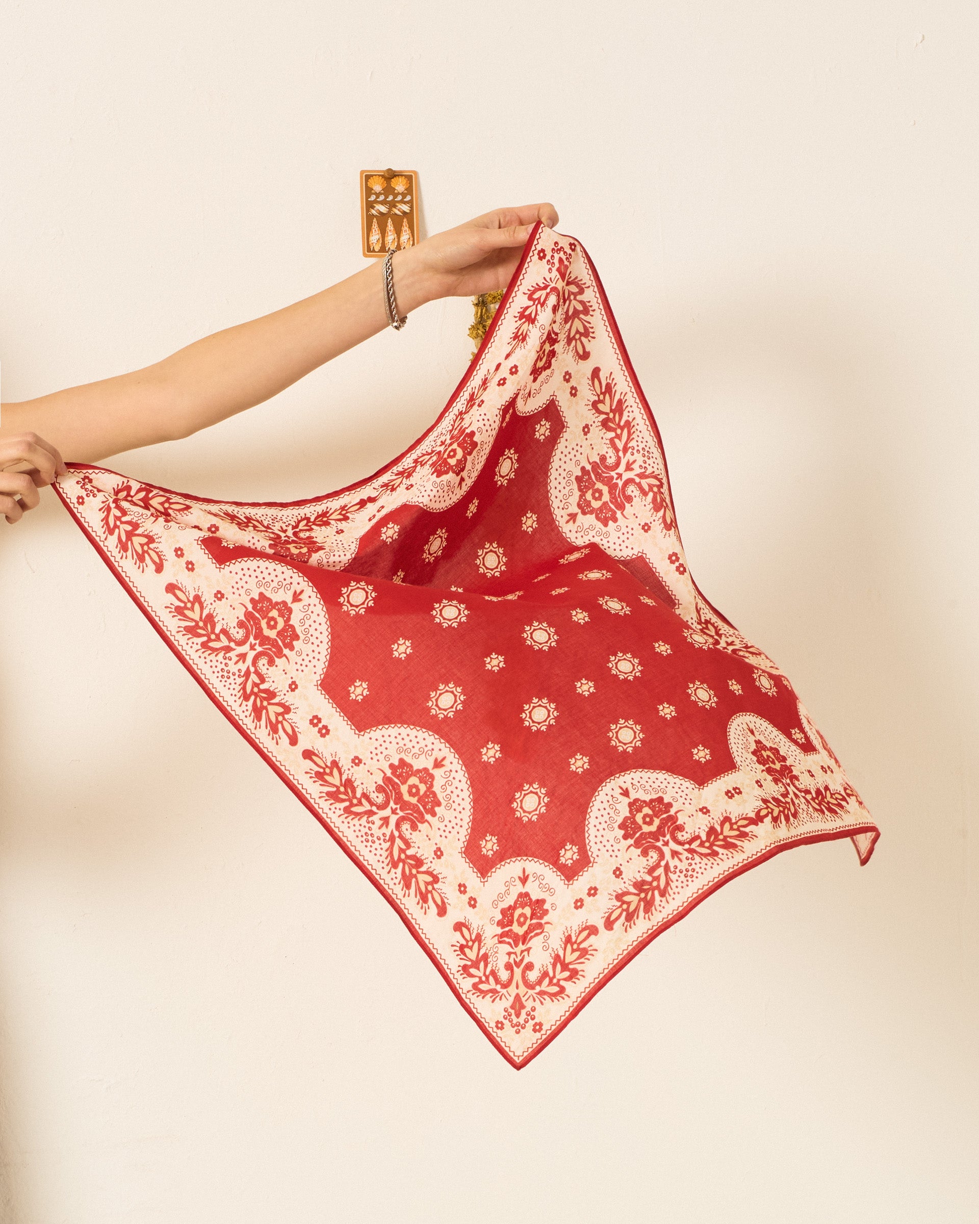 Someone holds up the Beau Lava Bandana, a vintage-inspired cotton accessory featuring a red and white pattern, against a plain light-colored wall.