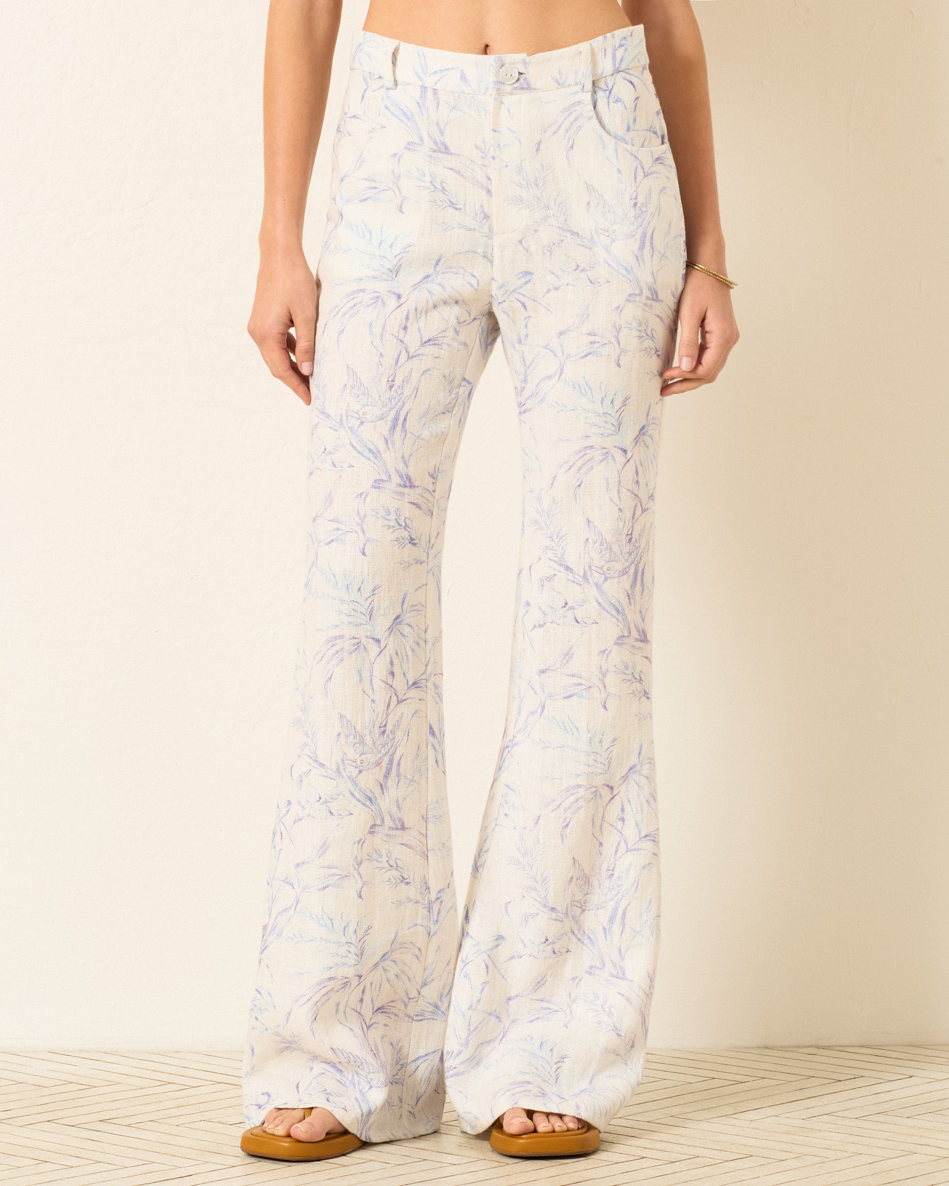A person wearing Bunny Avian Toile Jean—high-waisted, flared pants with a light pastel leaf pattern—stands on a patterned floor in sandals, showing a bare midriff.