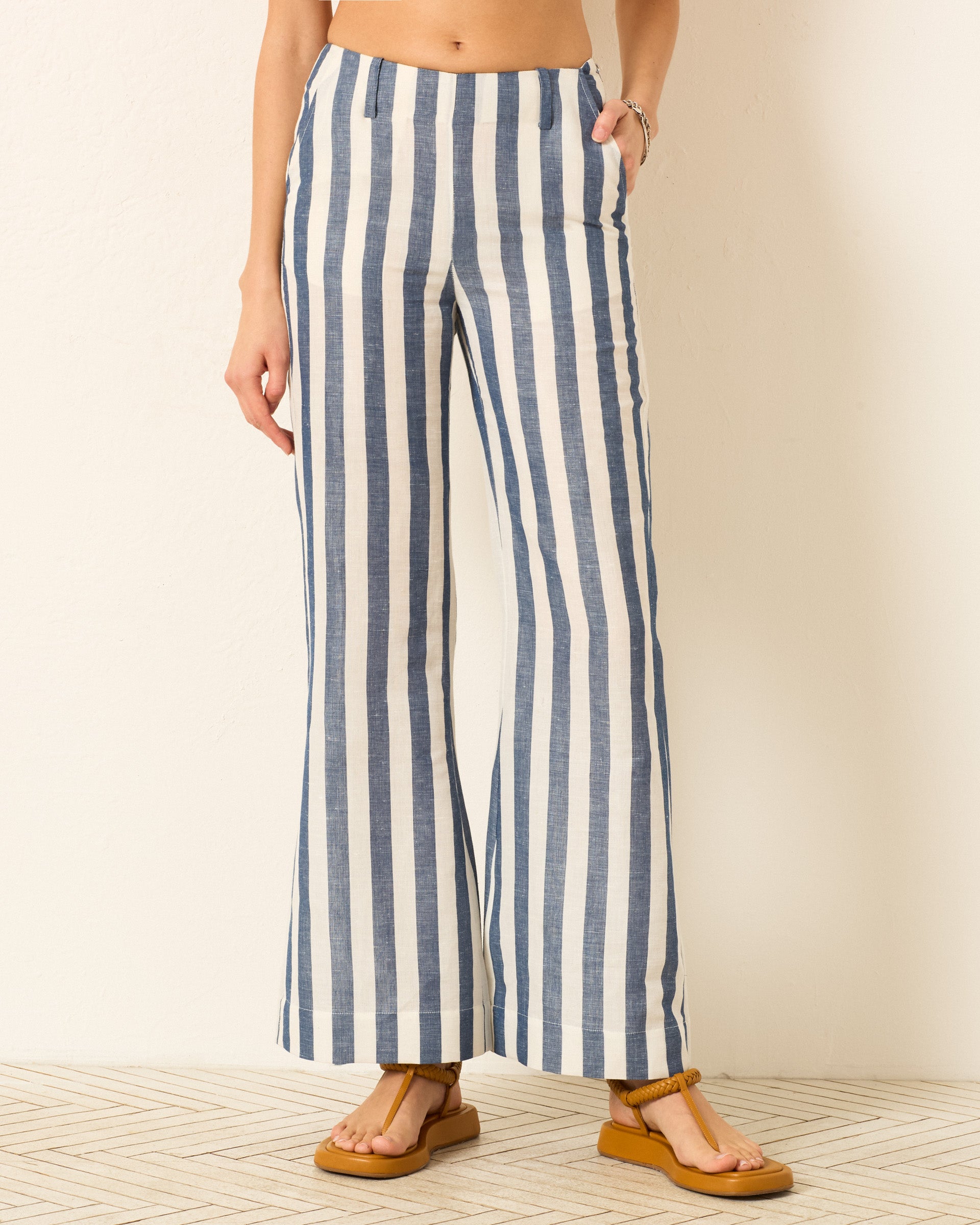 Embrace nautical style with the Charlie Sailor Stripe Pant—high-waisted, wide-leg linen pants in blue and white stripes—teamed with tan sandals for the perfect summer staple against a light wall.
