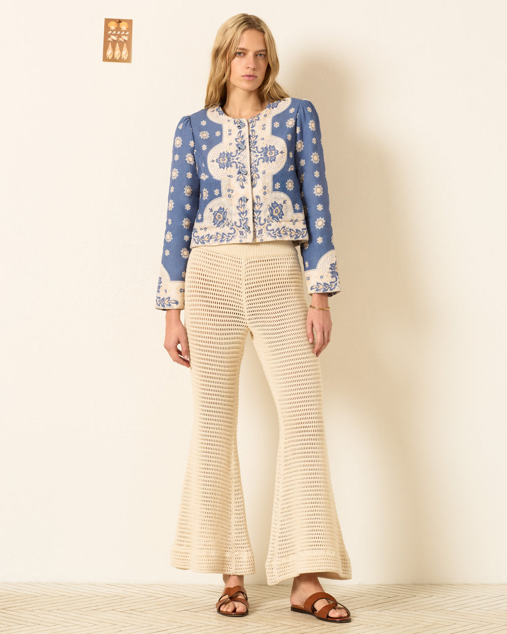 A woman stands against a light-colored wall wearing the Dorian Marina Bandana Jacket, featuring blue and white bandana print with handmade cowrie buttons, paired with cream crochet flared pants and brown sandals.