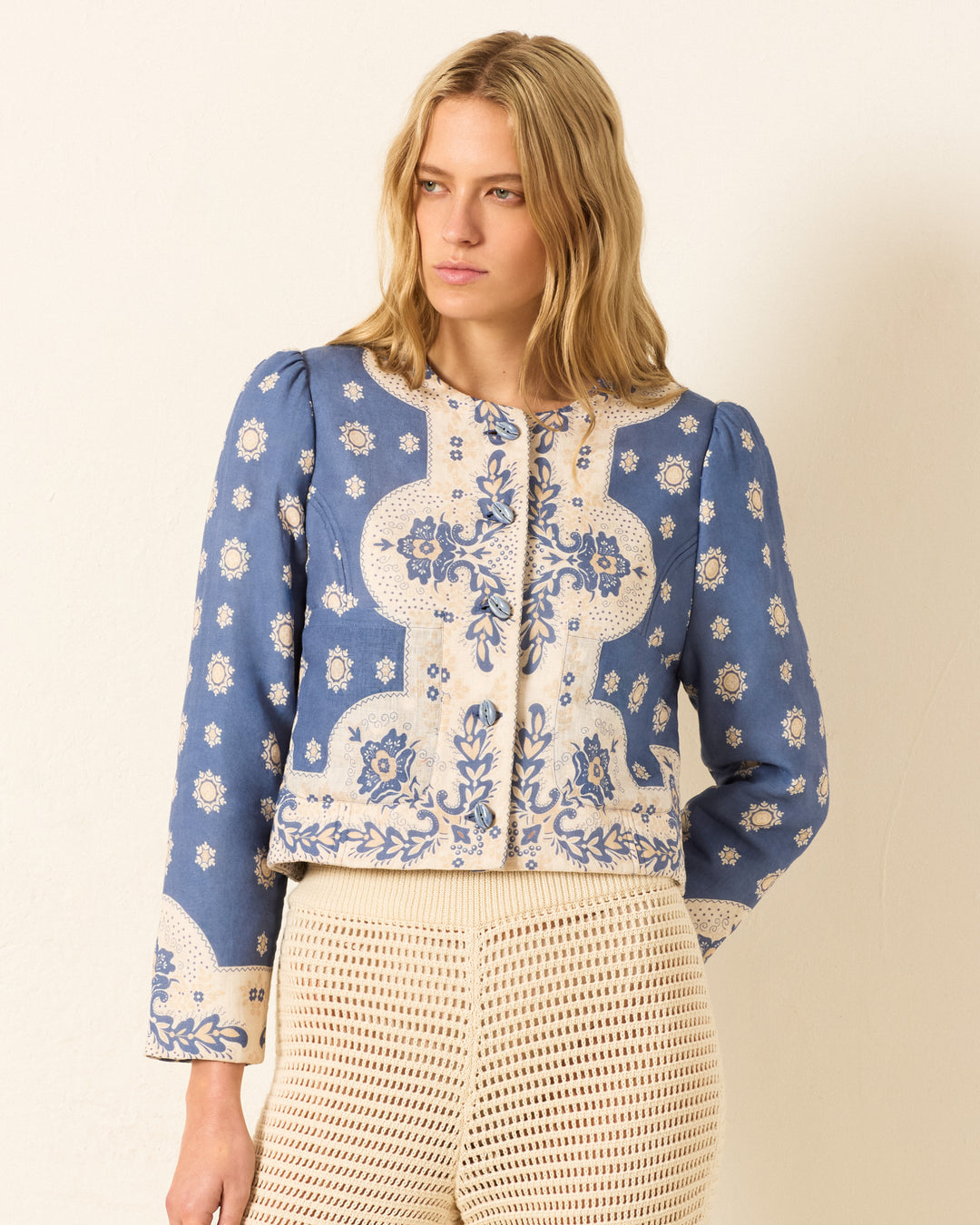 A woman with wavy blonde hair wears the Dorian Marina Bandana Jacket—blue and cream linen with floral patterns and handmade cowrie buttons—paired with cream crochet pants, standing against a plain light wall.
