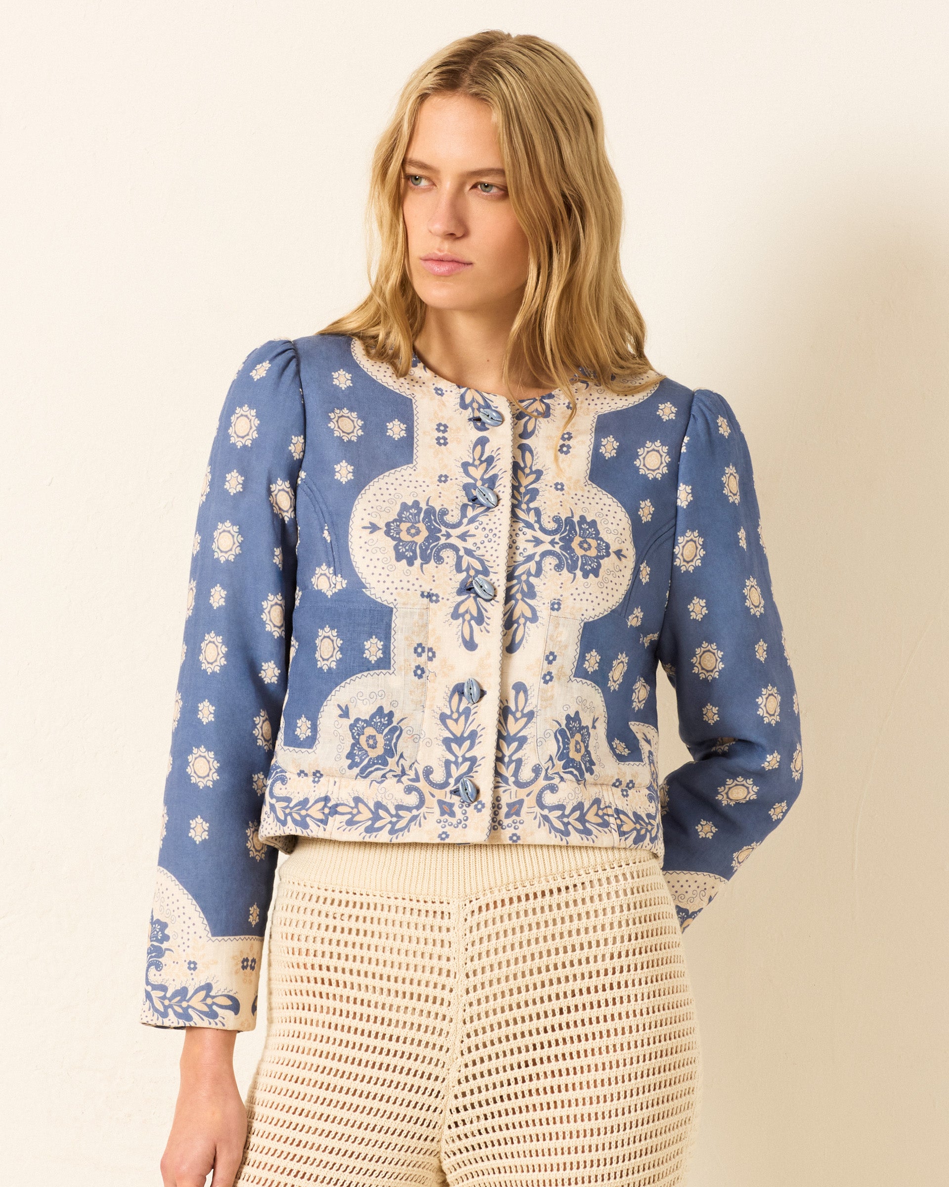 A woman with wavy blonde hair wears the Dorian Marina Bandana Jacket—blue and cream linen with floral patterns and handmade cowrie buttons—paired with cream crochet pants, standing against a plain light wall.