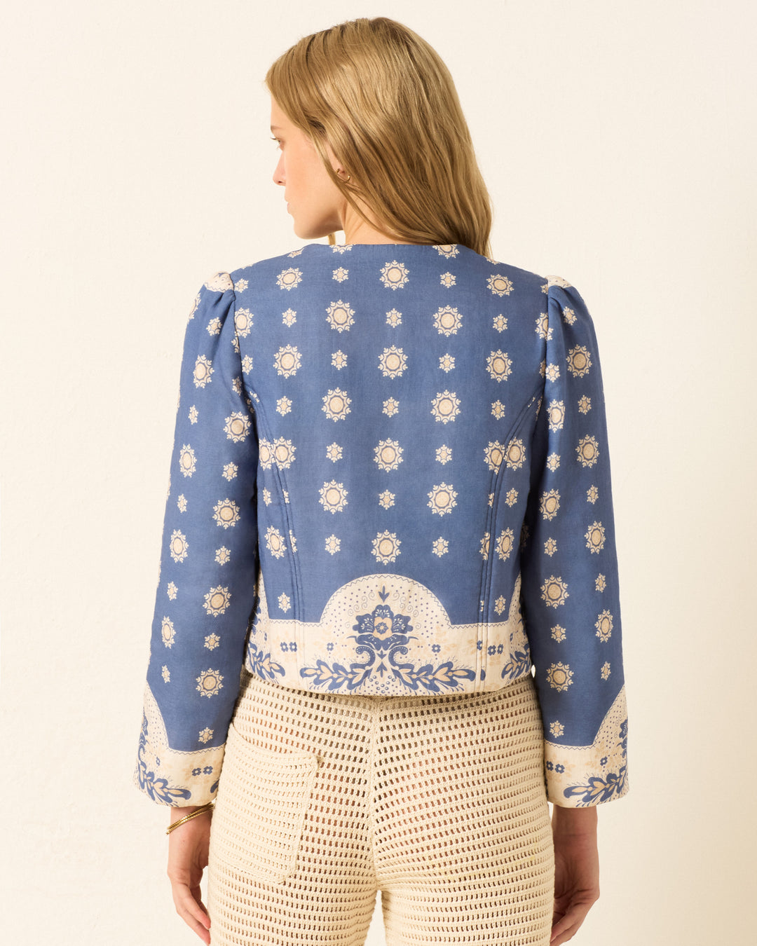 A woman stands facing away, wearing the Dorian Marina Bandana Jacket with cream floral designs and beige textured pants against a plain background.