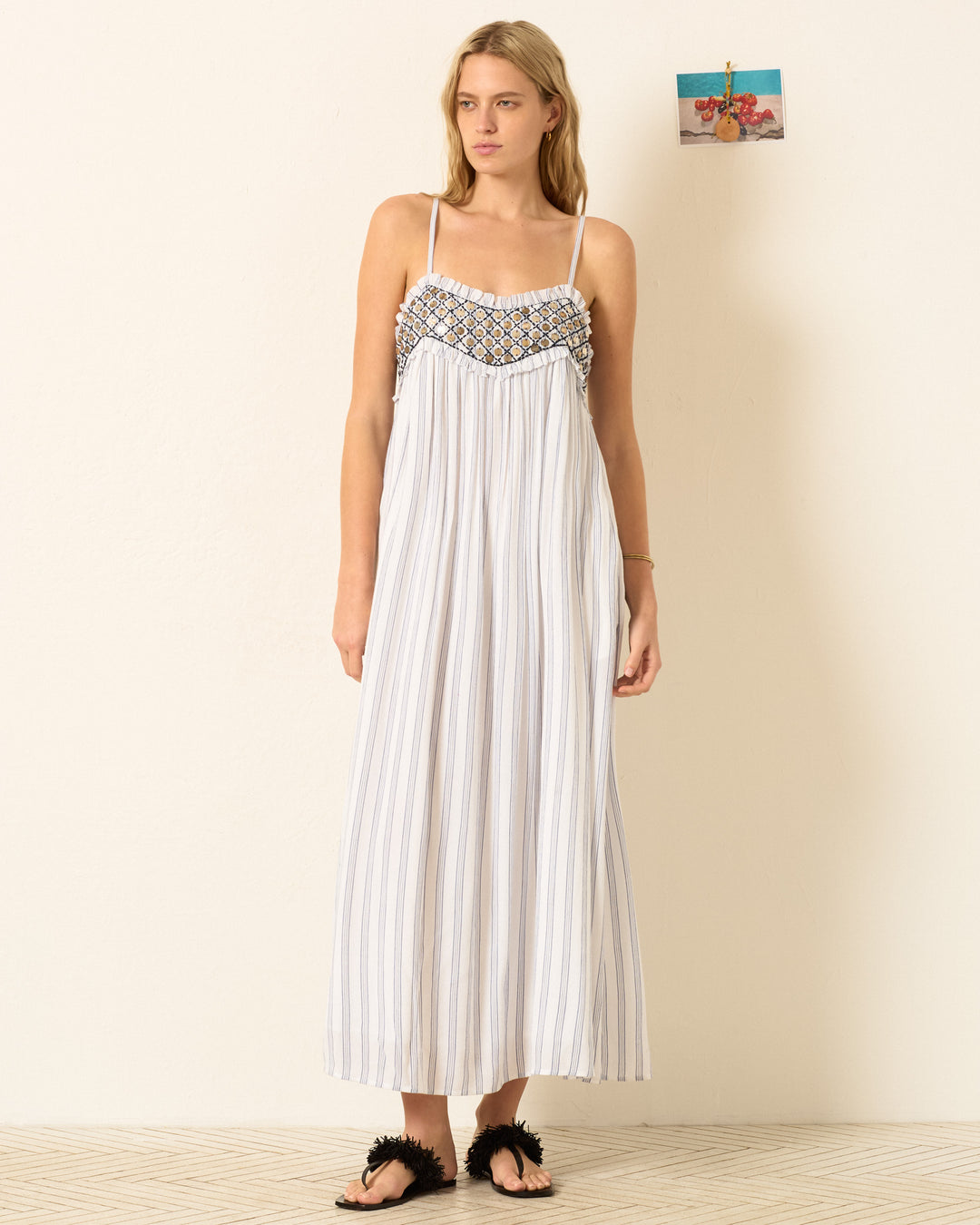 A woman stands indoors wearing the Eden Coastal Stripe Dress, featuring blue-and-white stripes and a patterned top, paired with black sandals. A small photo is pinned to the wall behind her.