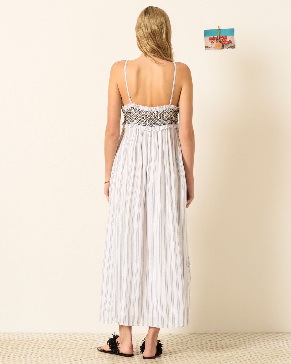 A woman with loose hair and sandals faces away, wearing the Eden Coastal Stripe Dress—a blue-and-white striped maxi dress with a smocked top. A small photo is pinned to the wall.