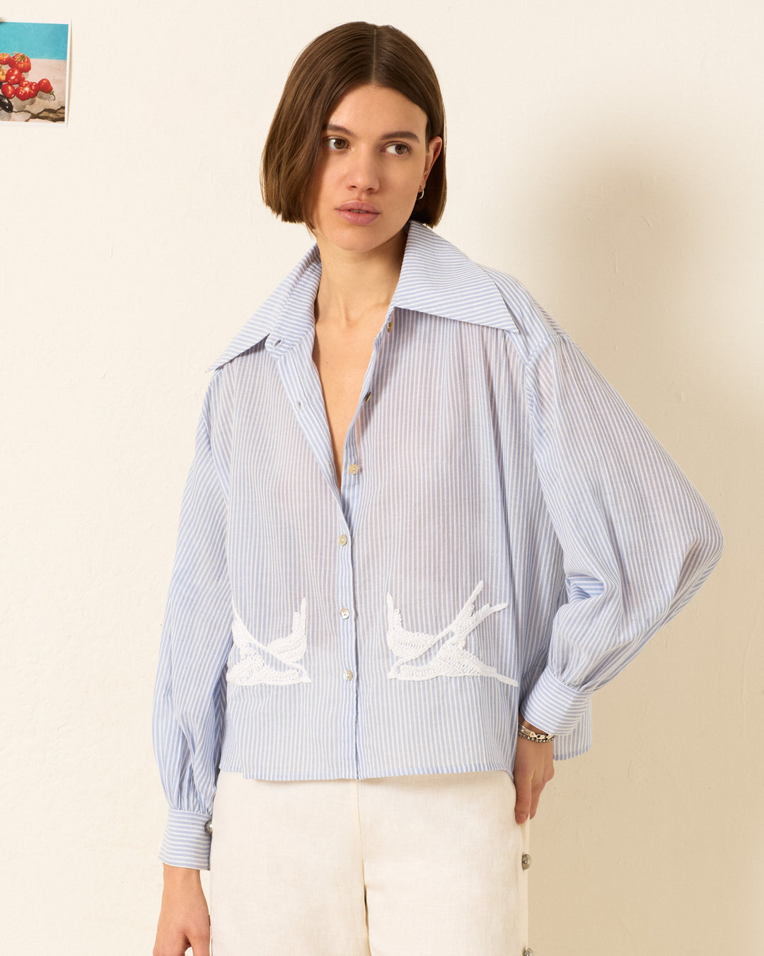 A woman stands against a plain wall wearing the Emery Swallow Stripe Shirt, which features blue-and-white stripes, a '70s-inspired dagger collar, and delicate white swallow embroidery.