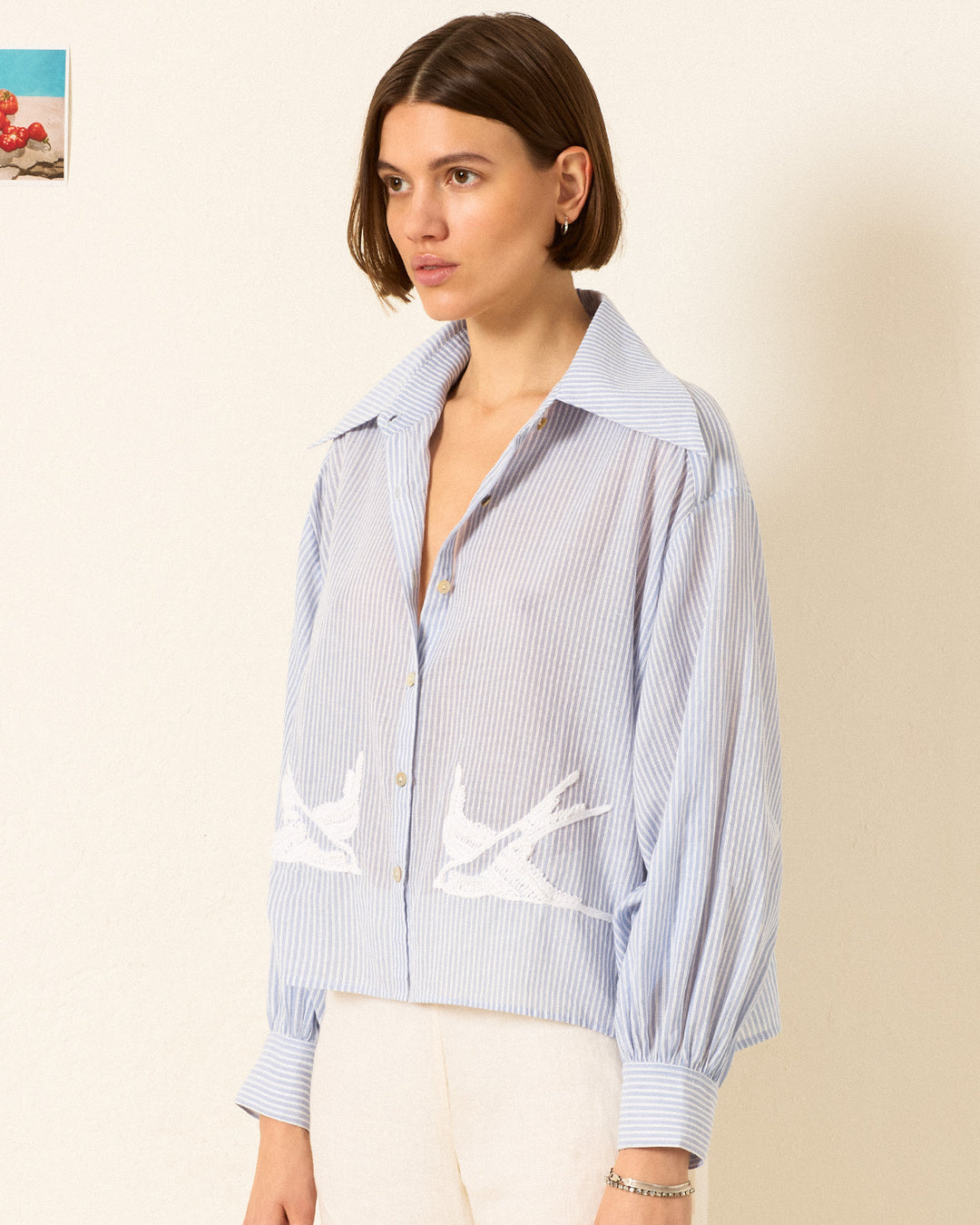 A woman wears the Emery Swallow Stripe Shirt—a blue-and-white striped cotton shirt with hand-embroidered bird motifs and a ’70s dagger collar—paired with white pants, standing against a plain wall.