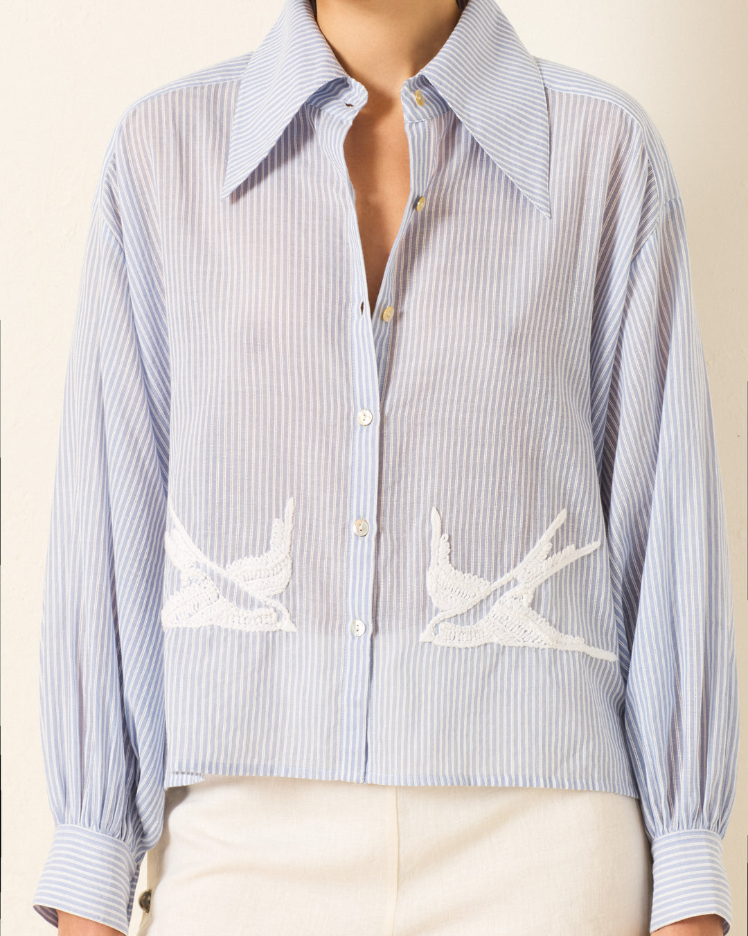 Wearing the Emery Swallow Stripe Shirt, which features blue-and-white stripes, a '70s-inspired dagger collar, sheer fabric, and white embroidered swallow designs near the hem.
