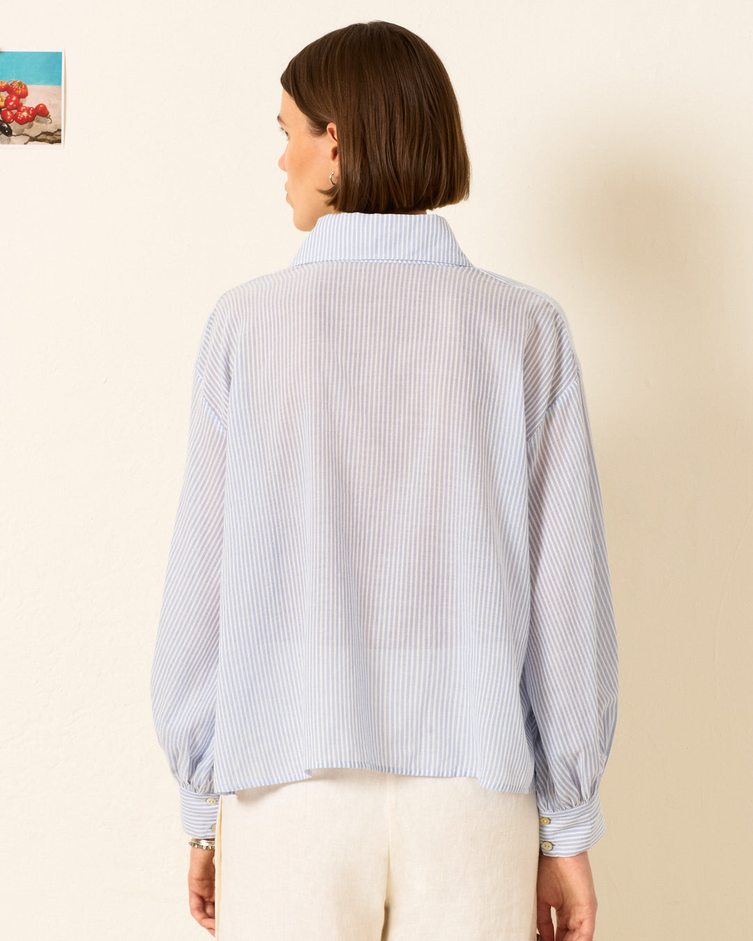A woman with short brown hair is shown from behind wearing the Emery Swallow Stripe Shirt, a loose blue-and-white striped long-sleeve top, paired with light-colored pants and standing against a plain wall.