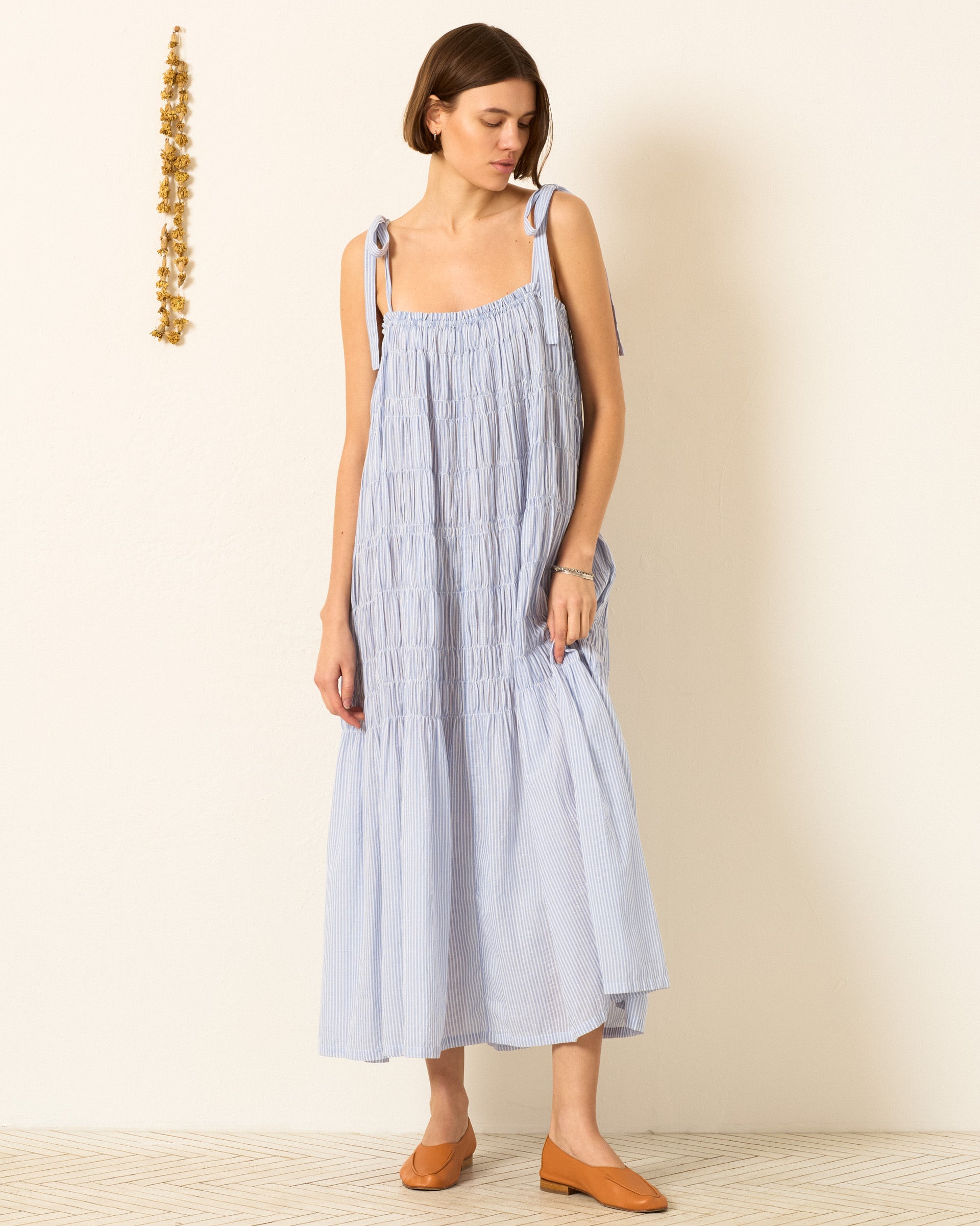 Wearing the Emrys Sky Stripe Dress—an airy, ankle-length, light blue dress with tie straps—a person stands against a plain, light wall in brown shoes, creating a subtle coastal vibe.