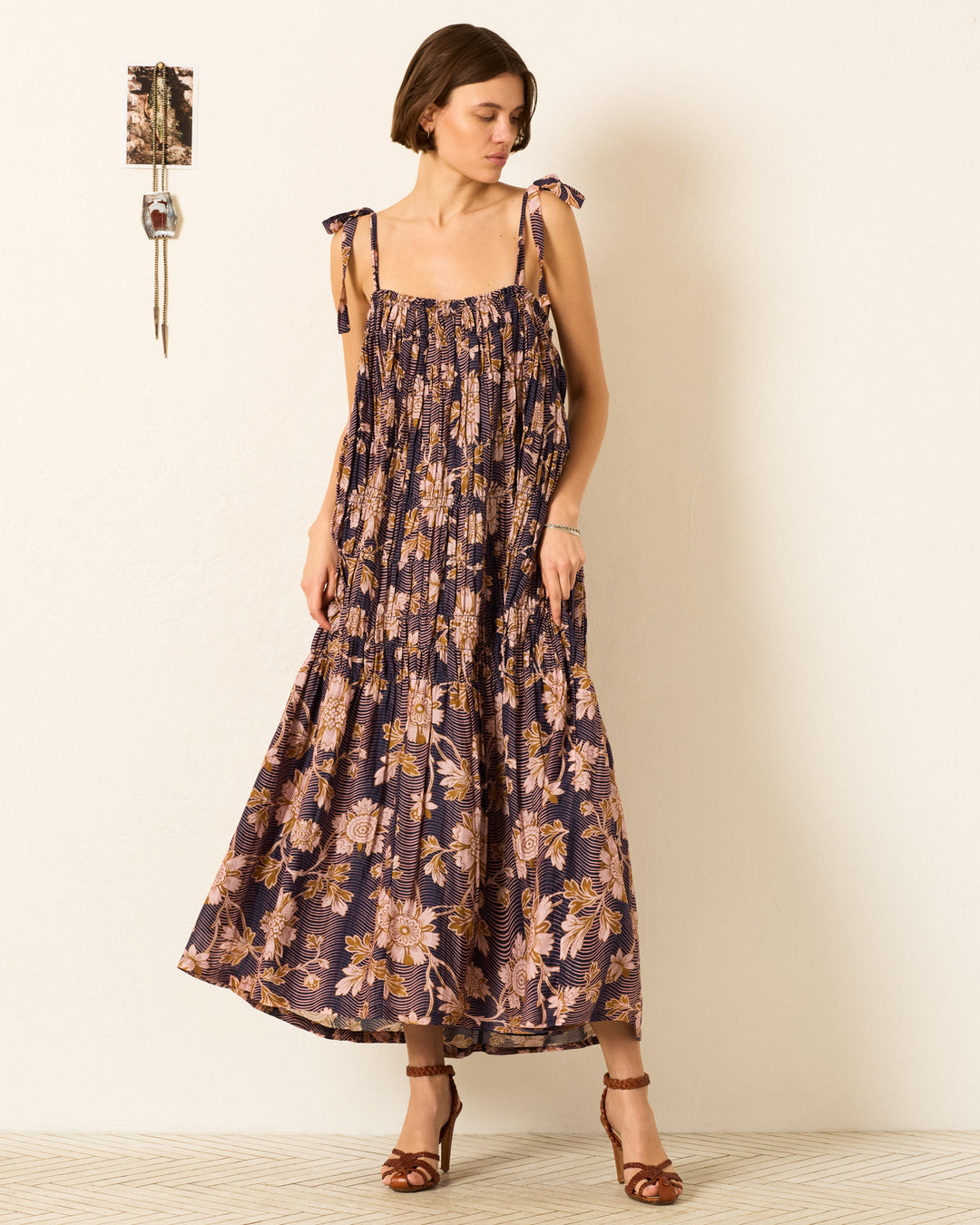 A woman stands indoors against a plain wall, wearing the Emrys Wavy Zinnia Dress—an ankle-length, relaxed maxi dress with a floral motif and shoulder ties. A small decorative item hangs beside her.