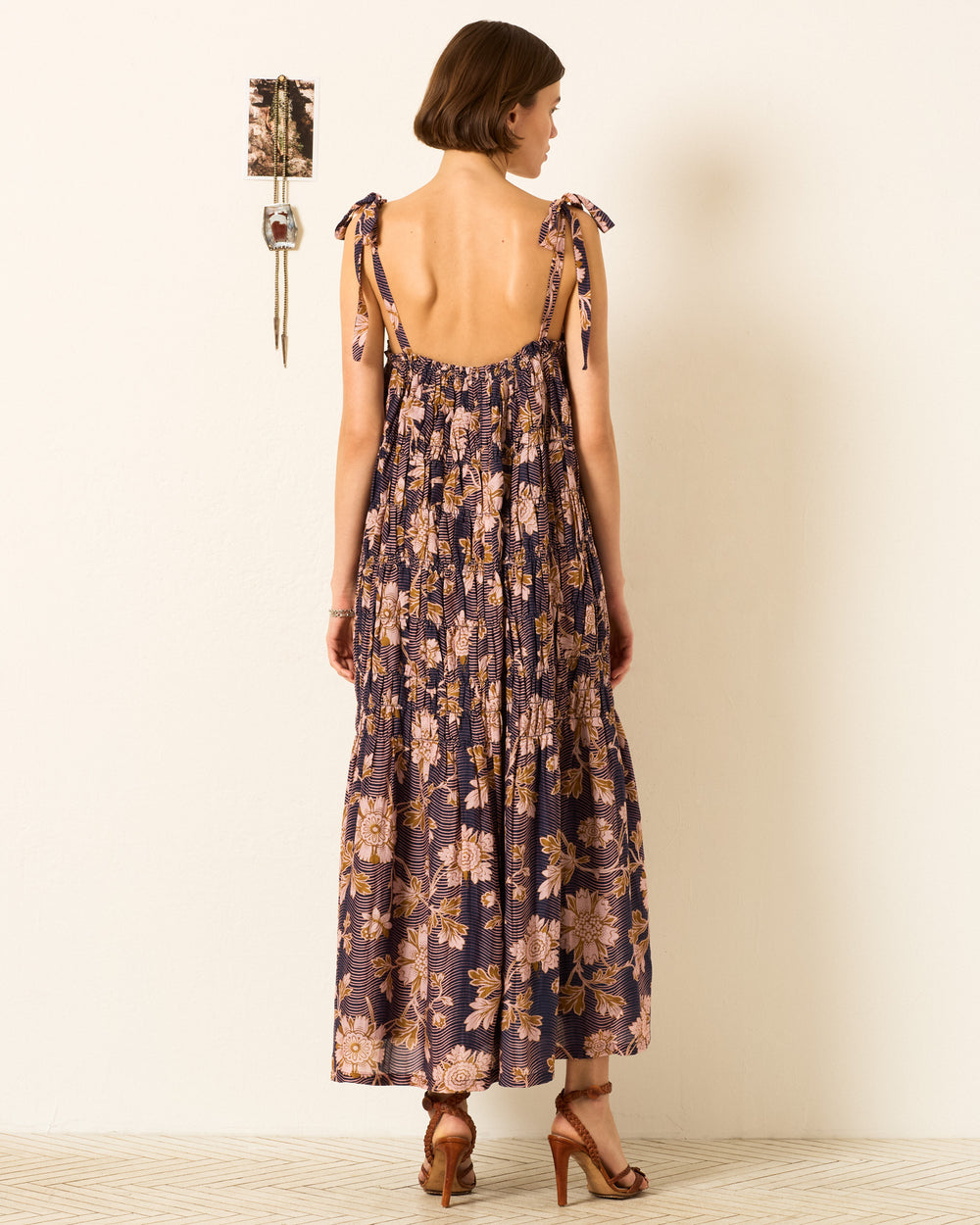 A woman stands indoors facing away, wearing the Emrys Wavy Zinnia Dress—a backless, sleeveless floral maxi with tie straps—paired with heeled sandals. A small wall hanging decor is visible beside her.