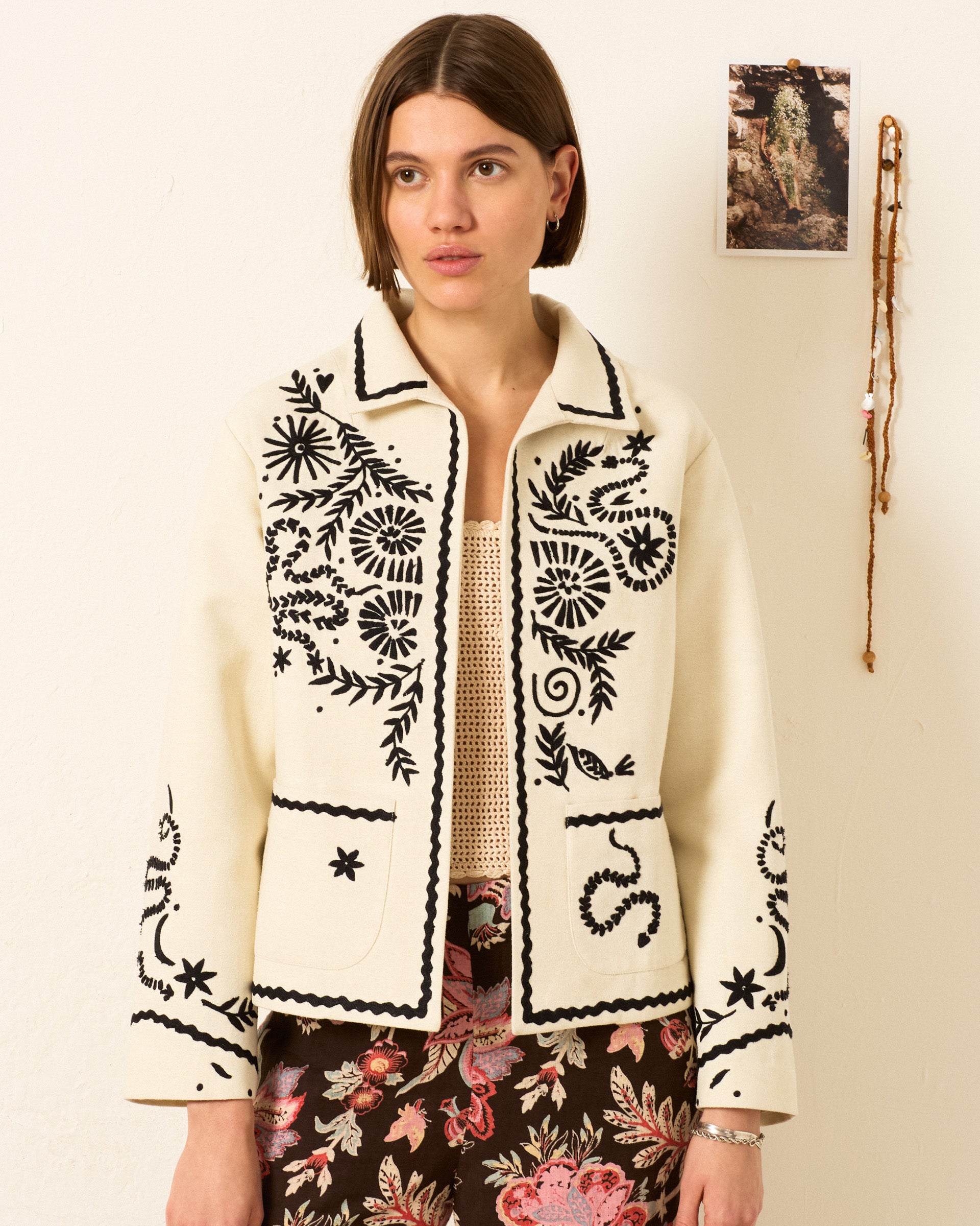 A woman with short brown hair wears the Francis Bone Snake Jacket, which is cream-colored with hand-embroidered black floral designs, paired with a patterned skirt, standing indoors near a wall decorated with a photo and hanging beads.