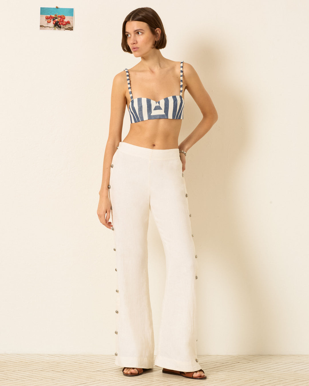 A woman stands indoors looking to the side, wearing a blue and white striped crop top and the Holland Pearl Linen Pant—high-waisted, white sailor pants with silver buttons. A small photo hangs on the wall behind her.