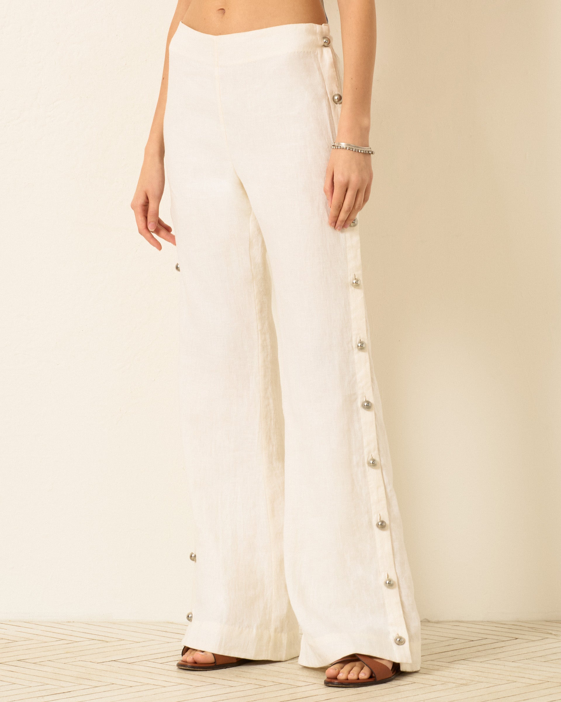 A person wears the Holland Pearl Linen Pant—high-waisted, white linen flared sailor pants with silver button detailing—while standing on a light patterned floor.