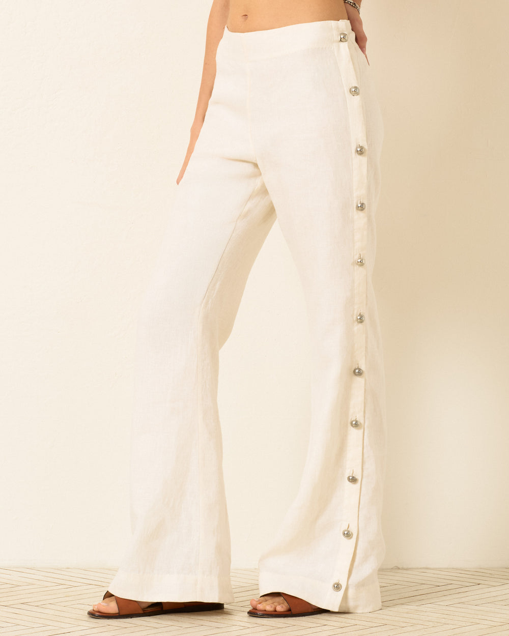 A person wearing Holland Pearl Linen Pant with silver buttons and brown sandals stands against a light background.
