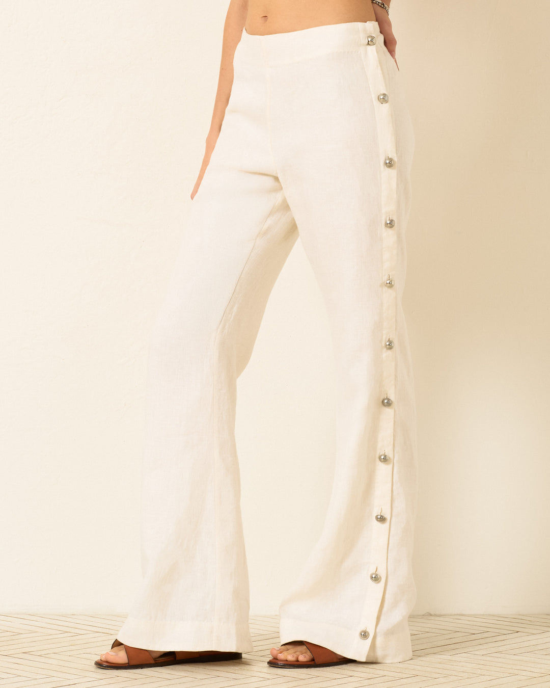A person wearing Holland Pearl Linen Pant with silver buttons and brown sandals stands against a light background.