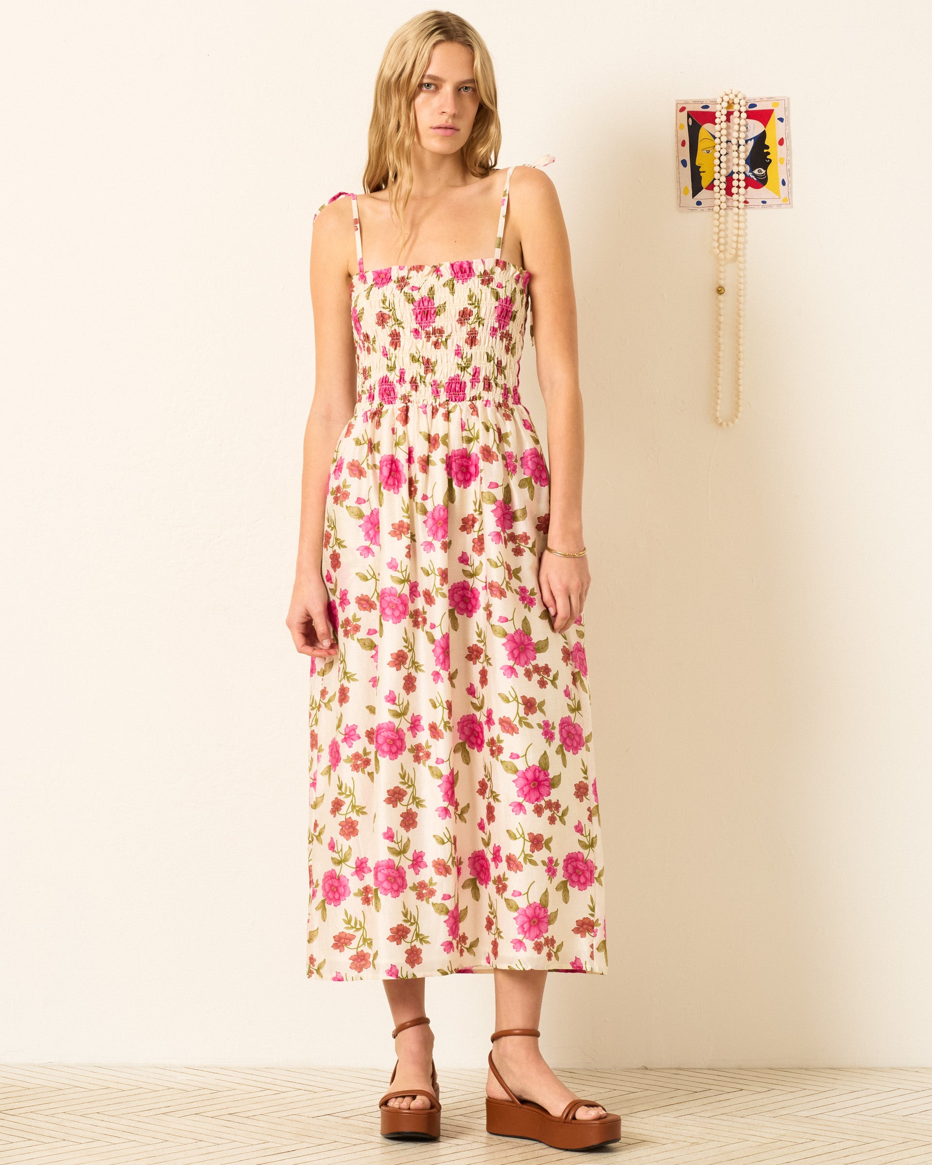 A woman stands against a plain wall, wearing the Kaia Pink Camellia Dress with a smocked bodice and pink floral print, paired with brown platform sandals. A small decorative piece hangs on the wall.