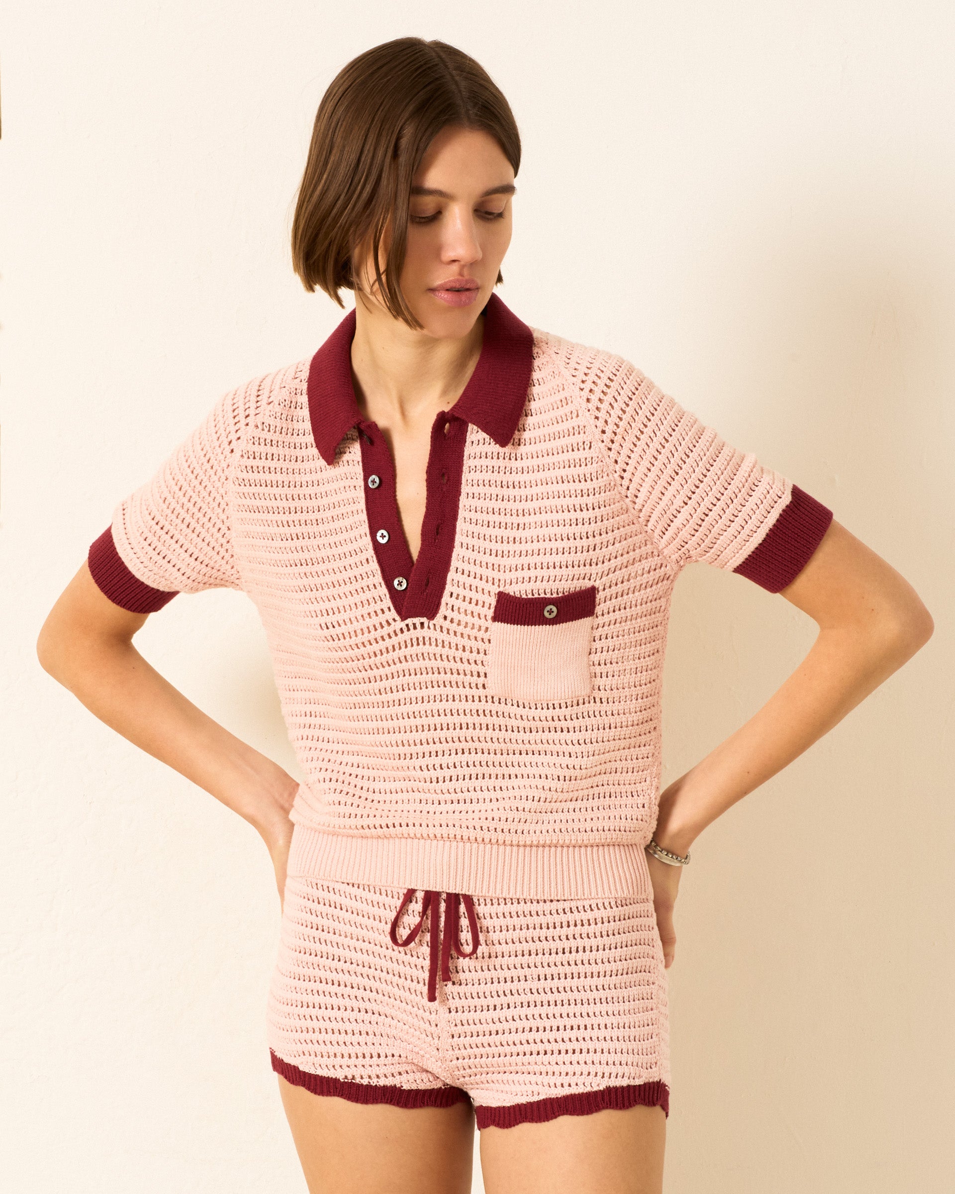 A woman wears the Lessi Blush Crochet Top, a light pink knit short-sleeve top with maroon trim and mother-of-pearl buttons, paired with matching shorts, standing against a plain light-colored wall.